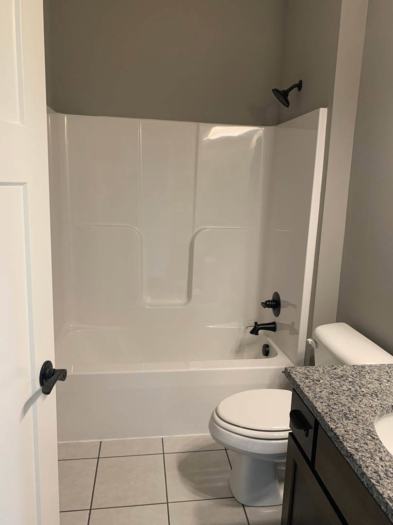 Modern bathroom with white bathtub and black fixtures, wall-mounted shower head, white toilet, stone countertop with undermount sink, and brushed metal door handle.