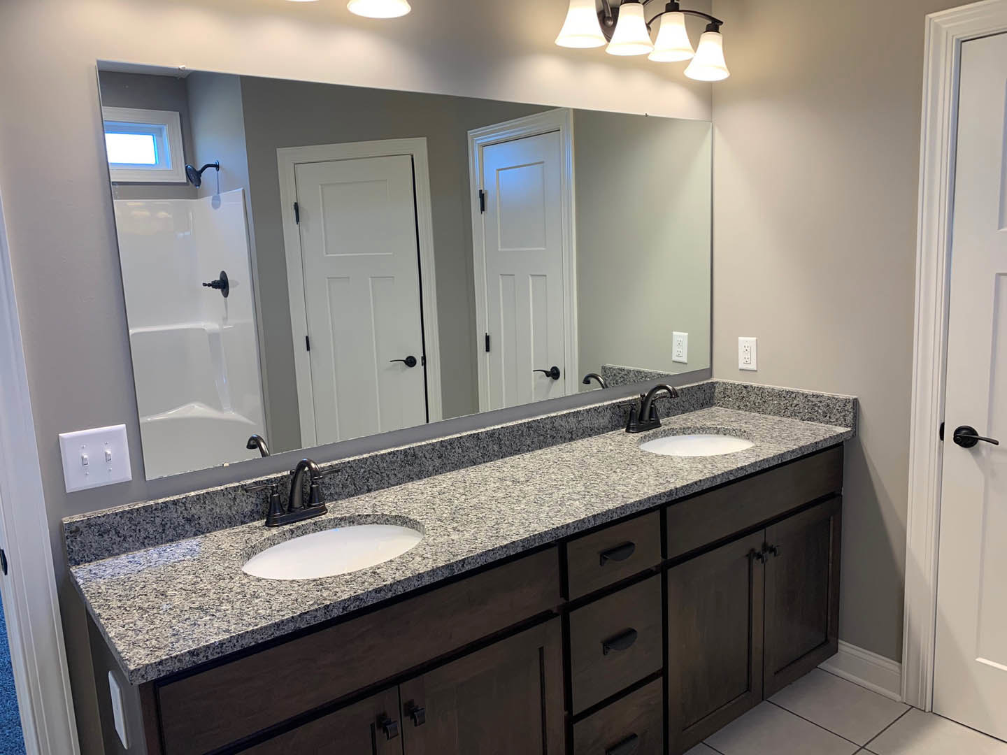 Bathroom with double sinks set in a granite countertop, large wall mirror above, white door with black handle, light switch on wall, and cabinetry beneath sinks
