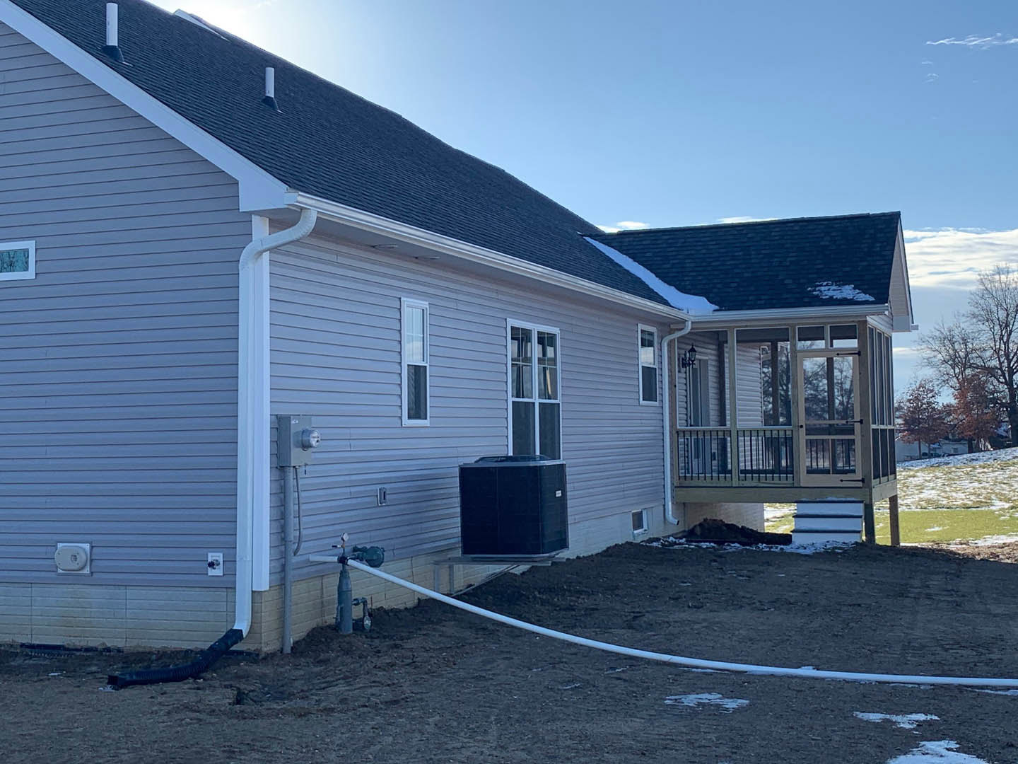 Modern home with gray siding, large windows, wooden deck, garden hose coiled near the foundation, and blue sky in the background