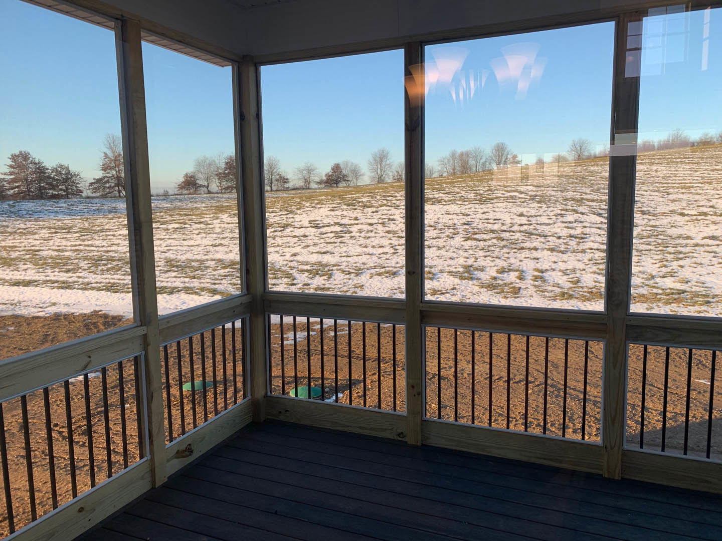 Dark wood flooring and wooden-framed windows overlooking a snowy field, deck with fence, blue sky, and distant trees
