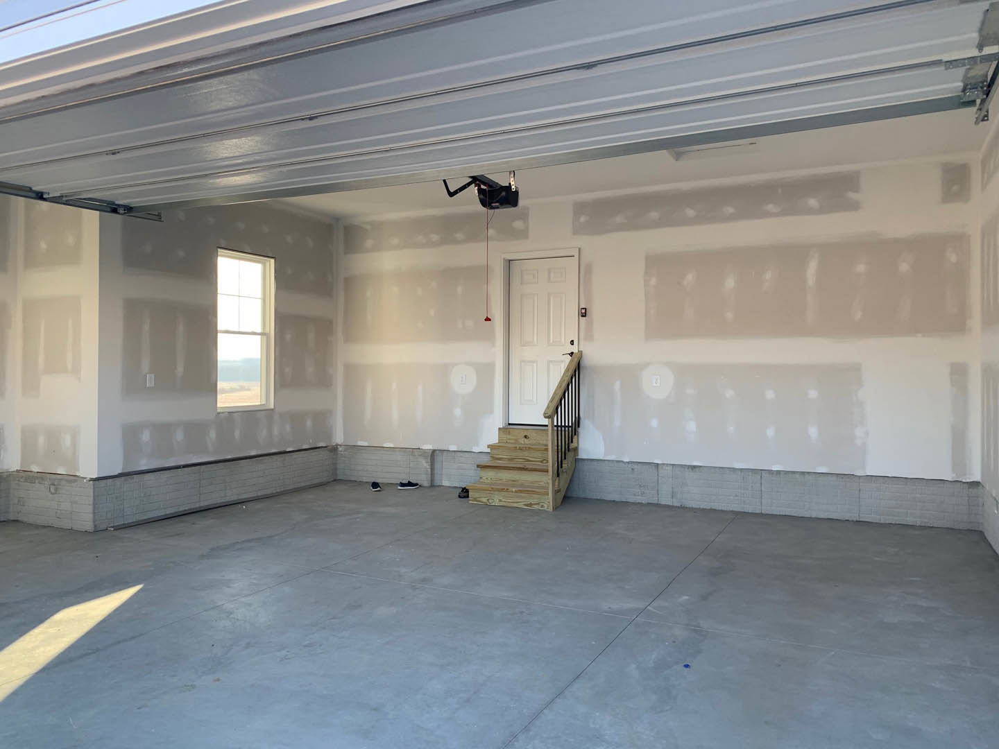 Concrete floor with wooden staircase featuring black railings, white door, white walls, rectangular window, and light switch.