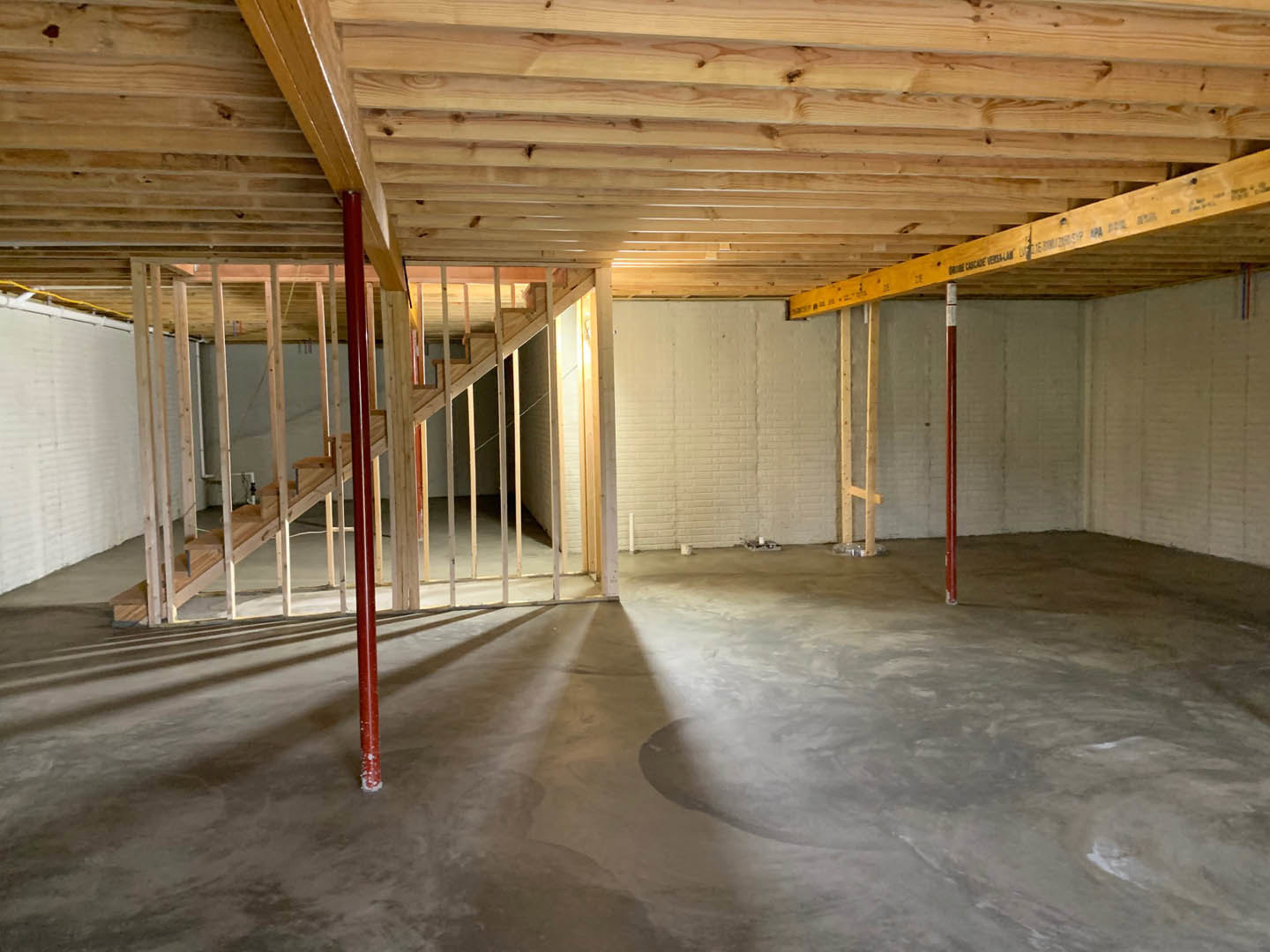 Open room with concrete floor, white painted wall, white brick accent wall, wooden ceiling with exposed beams, red metal support pole, and staircase with wooden steps