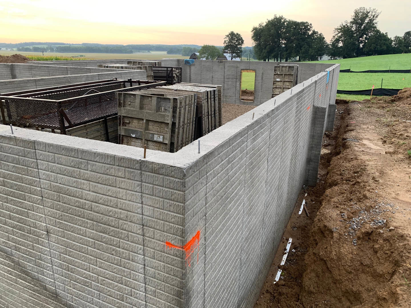 Concrete block wall with orange paint, adjacent to a dirt area featuring a large hole in the ground, outdoor setting with trees and sky visible in the background.
