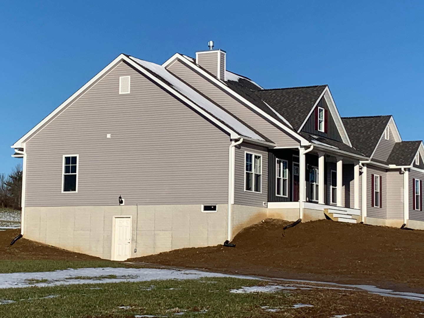 Two-story house with gray siding, white trim, black-framed windows, and a white front door, set on a grassy lawn with puddles, backed by a wooded hill under a cloudy sky