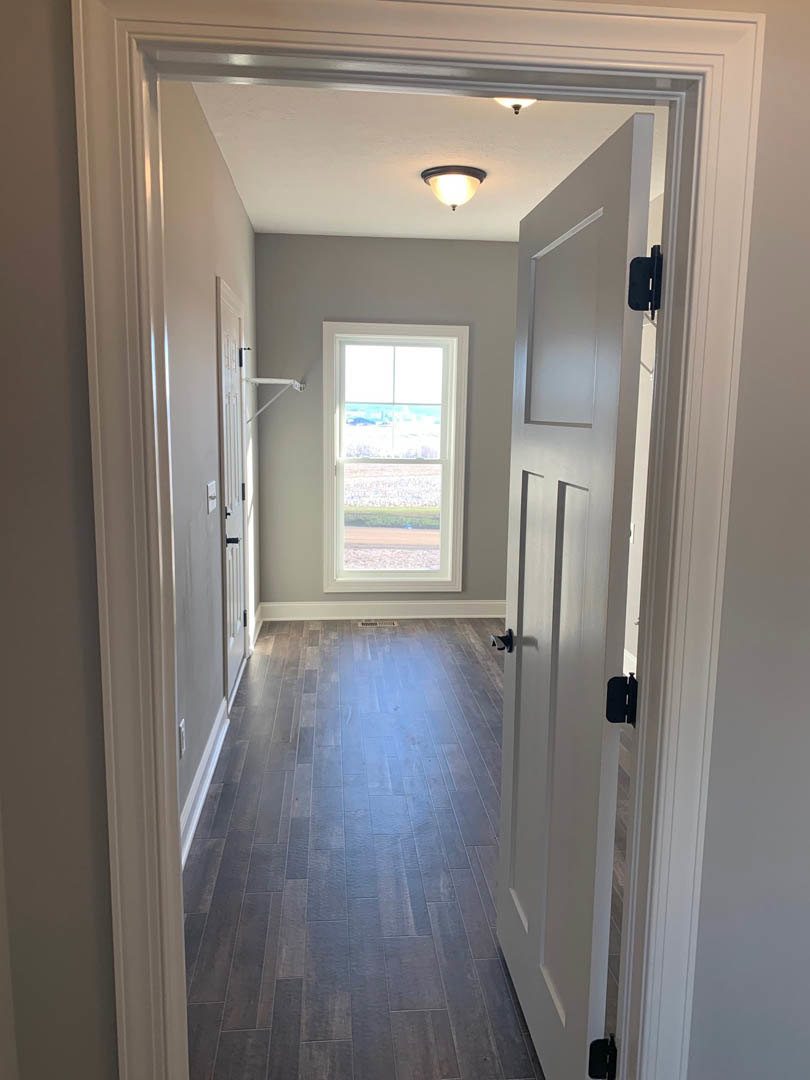Hallway with wide plank wood flooring, white walls, ceiling light fixture, window overlooking a grassy field, white door with black handle