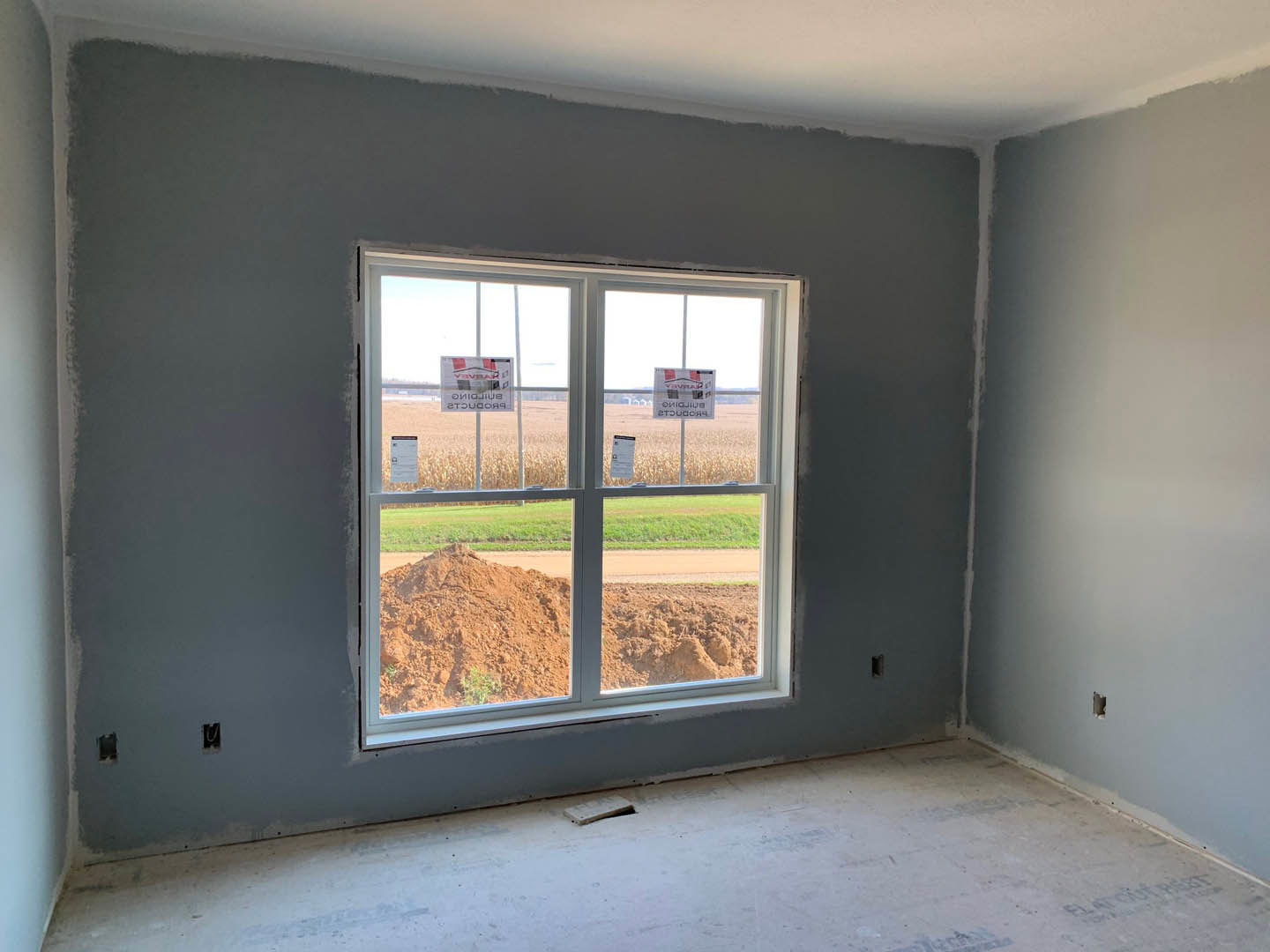 Unfinished room with plaster walls, window facing a pile of dirt outside, exposed subfloor with a hole, and construction signs taped to the glass
