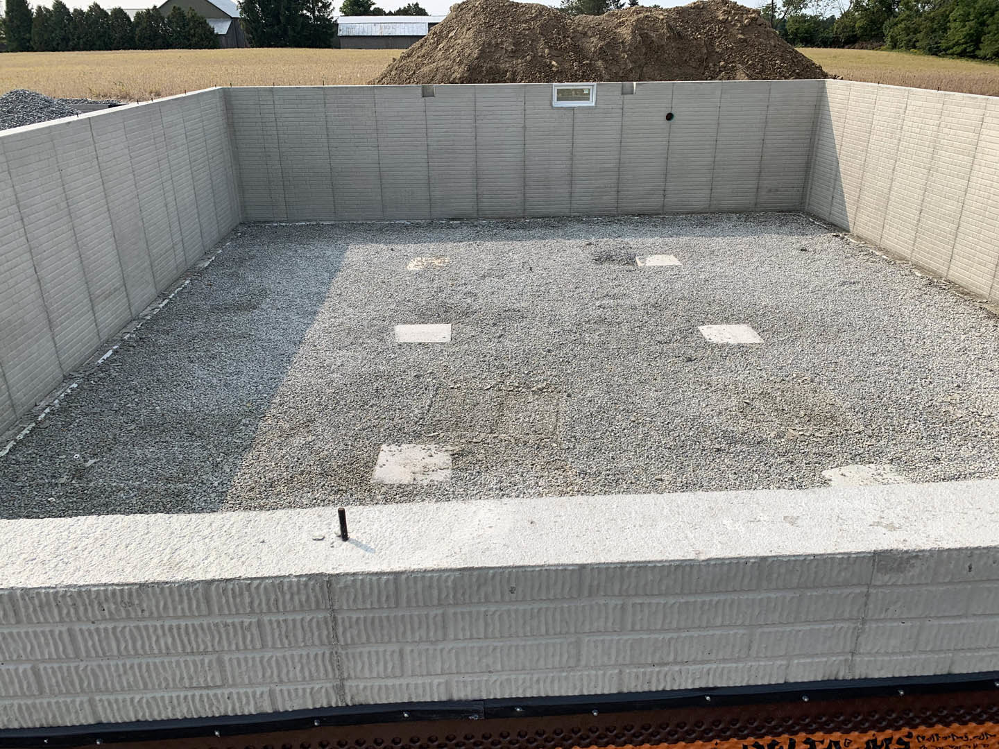Concrete foundation surrounded by gravel and dirt, with a pile of earth and trees in the background