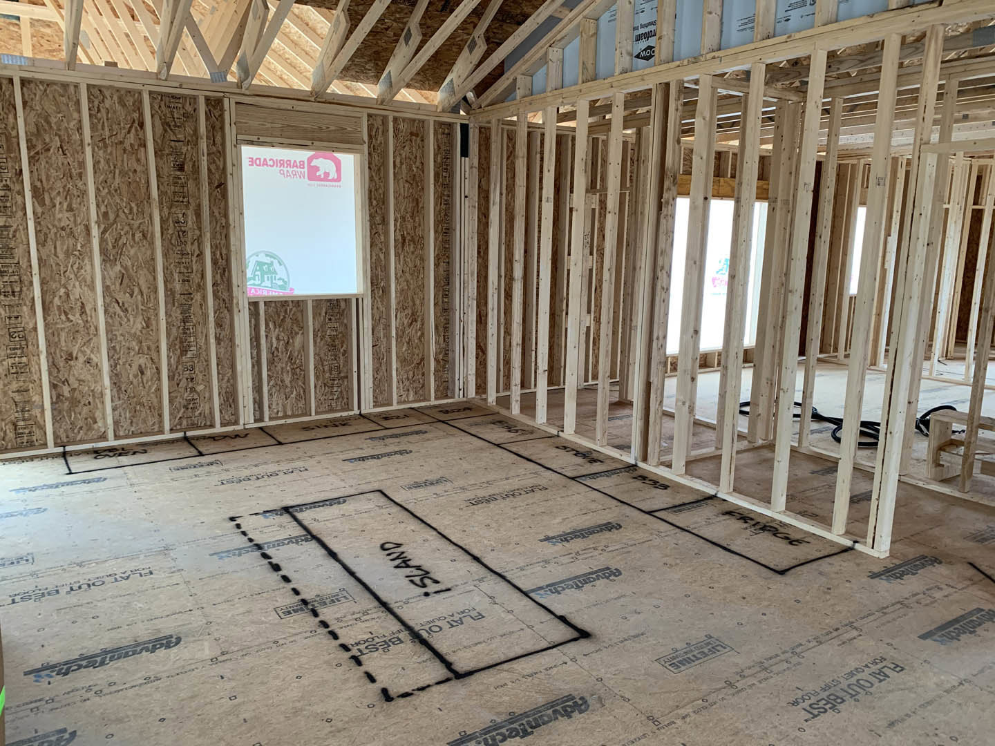 Room under construction with exposed wood framing, unfinished floor marked with black lines, large window featuring a pink logo on a white board.