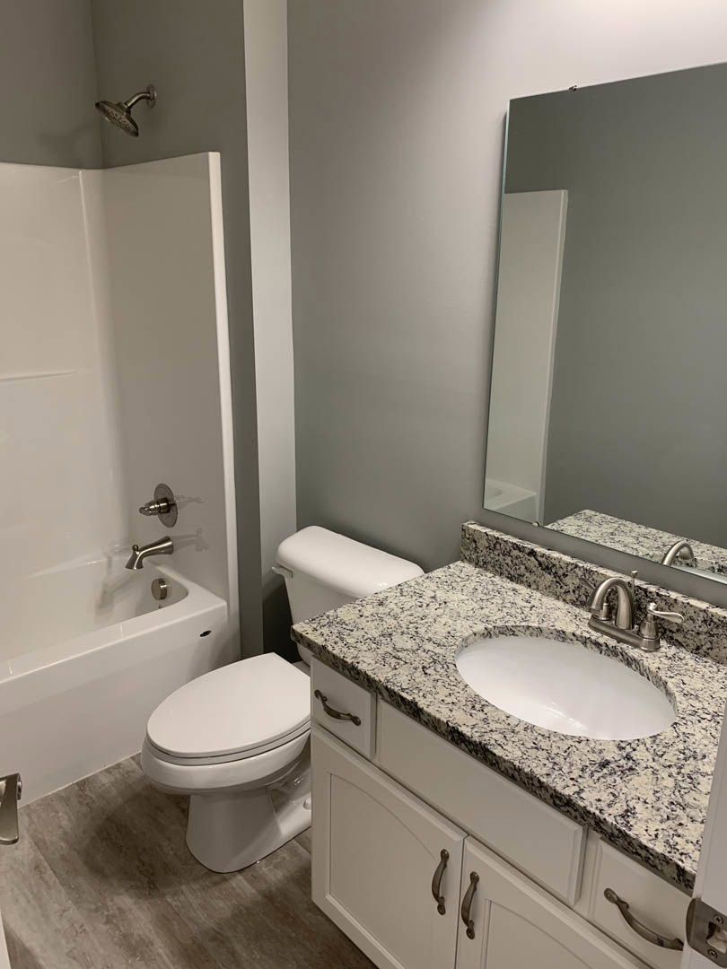 Marble countertop vanity with white sink and silver faucet, wall-mounted mirror, white toilet, tiled floor, partial view of bathtub