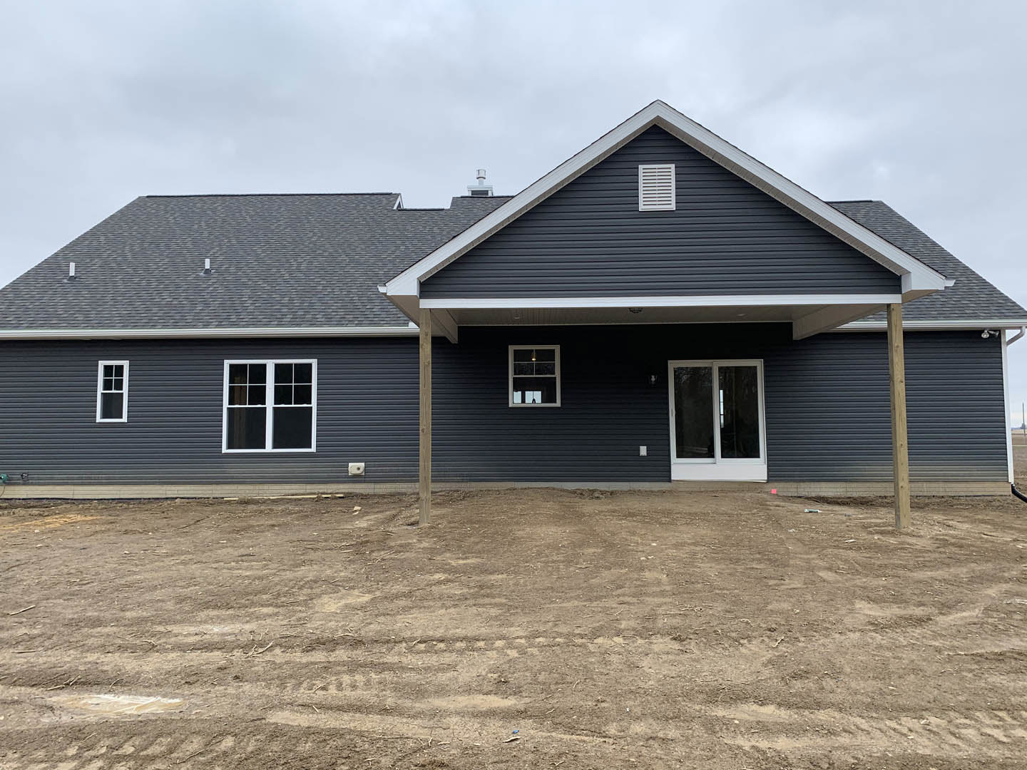 Single-story house with light-colored siding, white-framed windows, exterior wall vent, dirt yard featuring tire tracks, illuminated interior visible through one window, clear sky