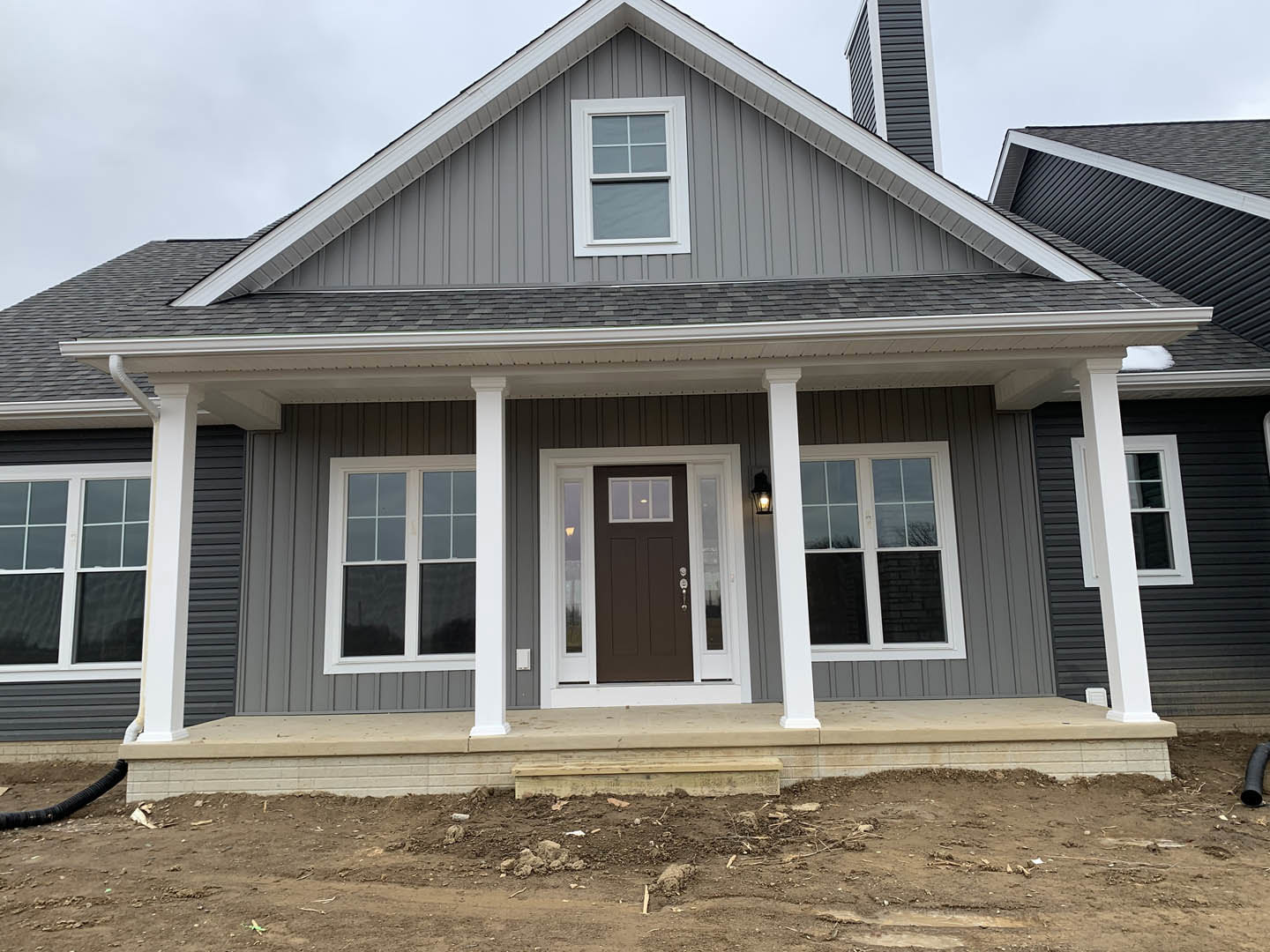 Brown front door with glass panes, flanked by white pillars on a porch, white siding exterior, sash window, wooden steps, dirt and debris near entry.
