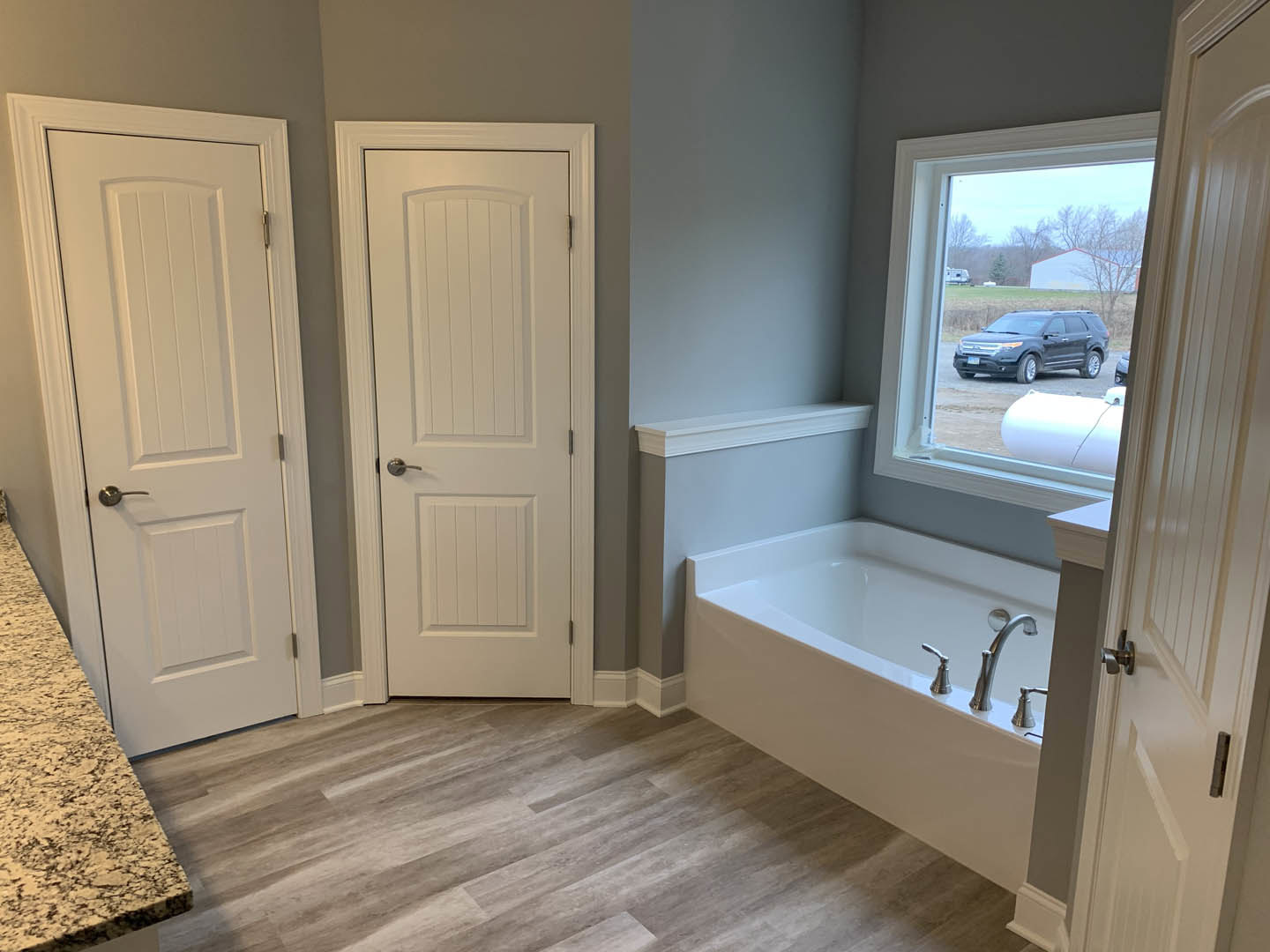 Freestanding bathtub with chrome faucets on wood floor, large window with white trim, light-colored walls, and built-in cabinetry