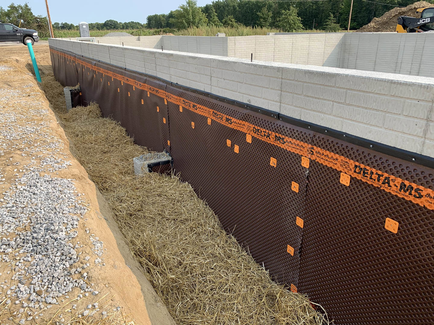 Brown and orange plastic sheeting covering an exterior wall, with straw and rocks piled nearby, a blue pole in the dirt, and a concrete drainage system visible in the foreground.