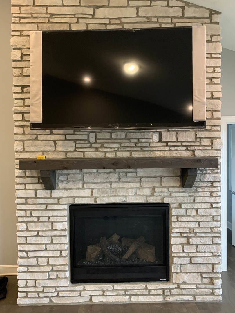 TV mounted above a brick fireplace with stacked wood logs in the hearth, wooden mantel shelf, and exposed wood beam with visible knot.