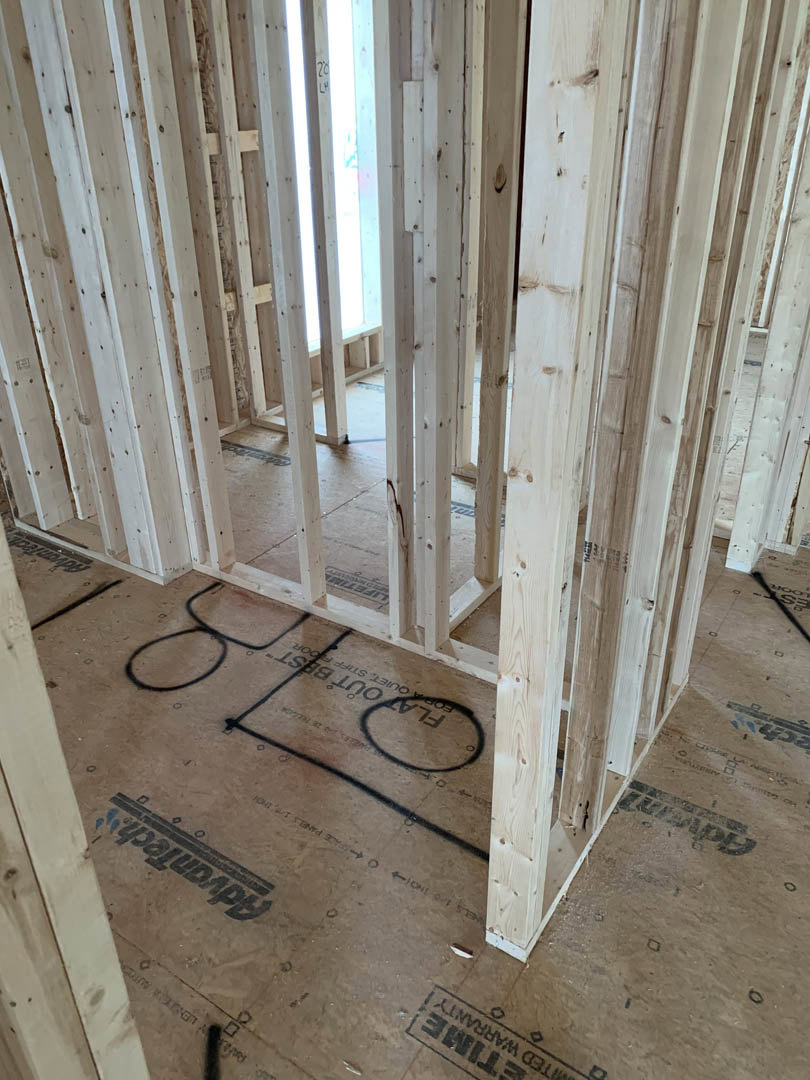 Exposed wood framing and subfloor inside a house under construction, with visible insulation, unfinished plaster, and construction markings on the floor