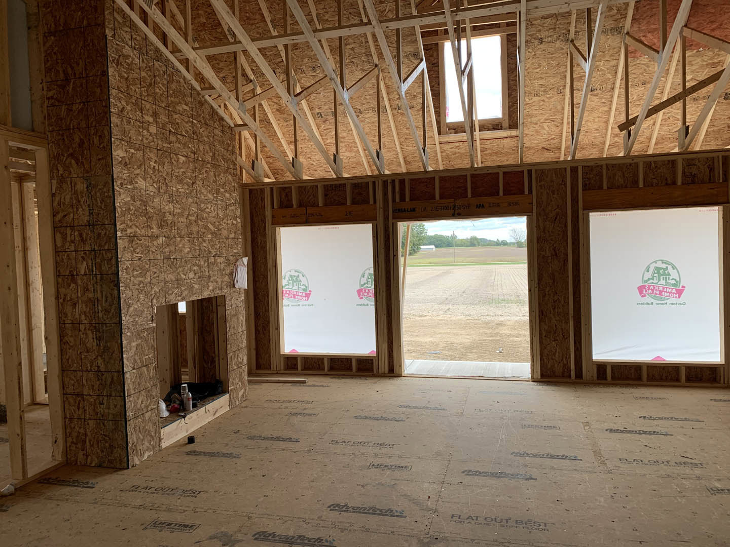 Spacious room with large windows, white walls featuring green and pink logos, light-colored flooring, and partial view of a dirt field and trees outside