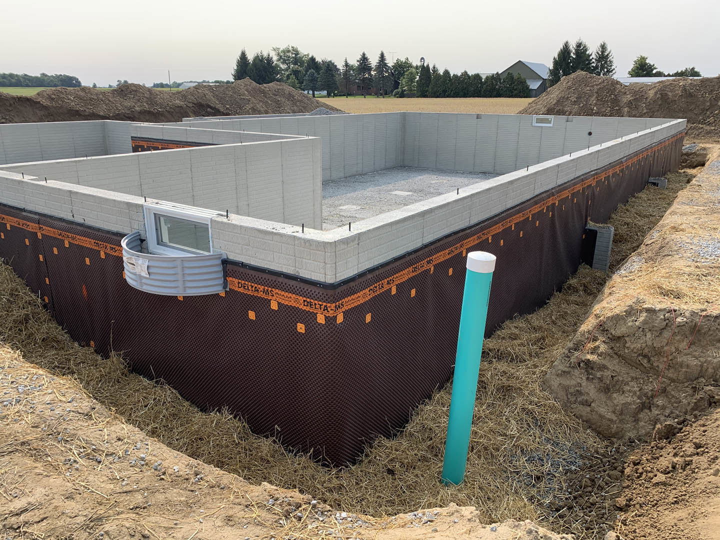 Concrete house foundation with exposed blue pipe, hay covering ground, window displaying white sign, construction materials scattered, row of leafy trees in background under clear
