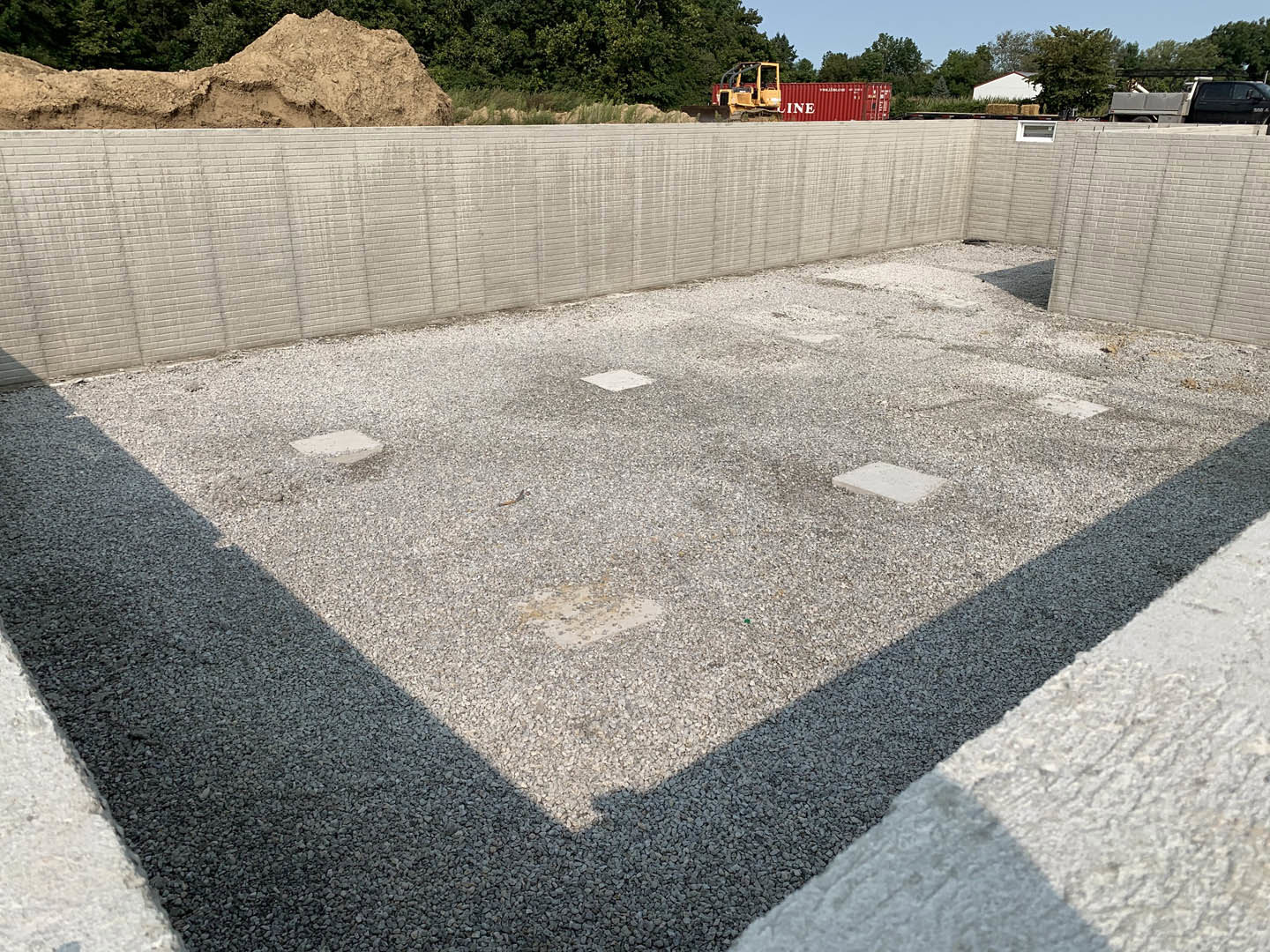Concrete foundation surrounded by a pile of gravel, yellow forklift near containers, gravel road with square stones, white rectangular object on grey surface, dirt hill in