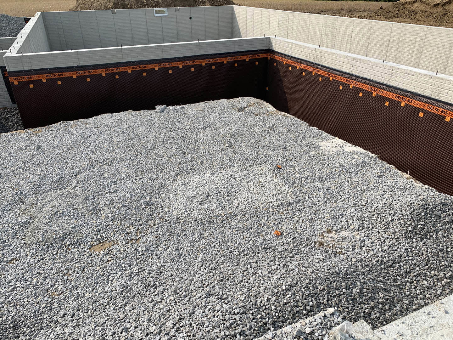 Pile of gray gravel beside a concrete wall with black and orange tape marking the foundation edge