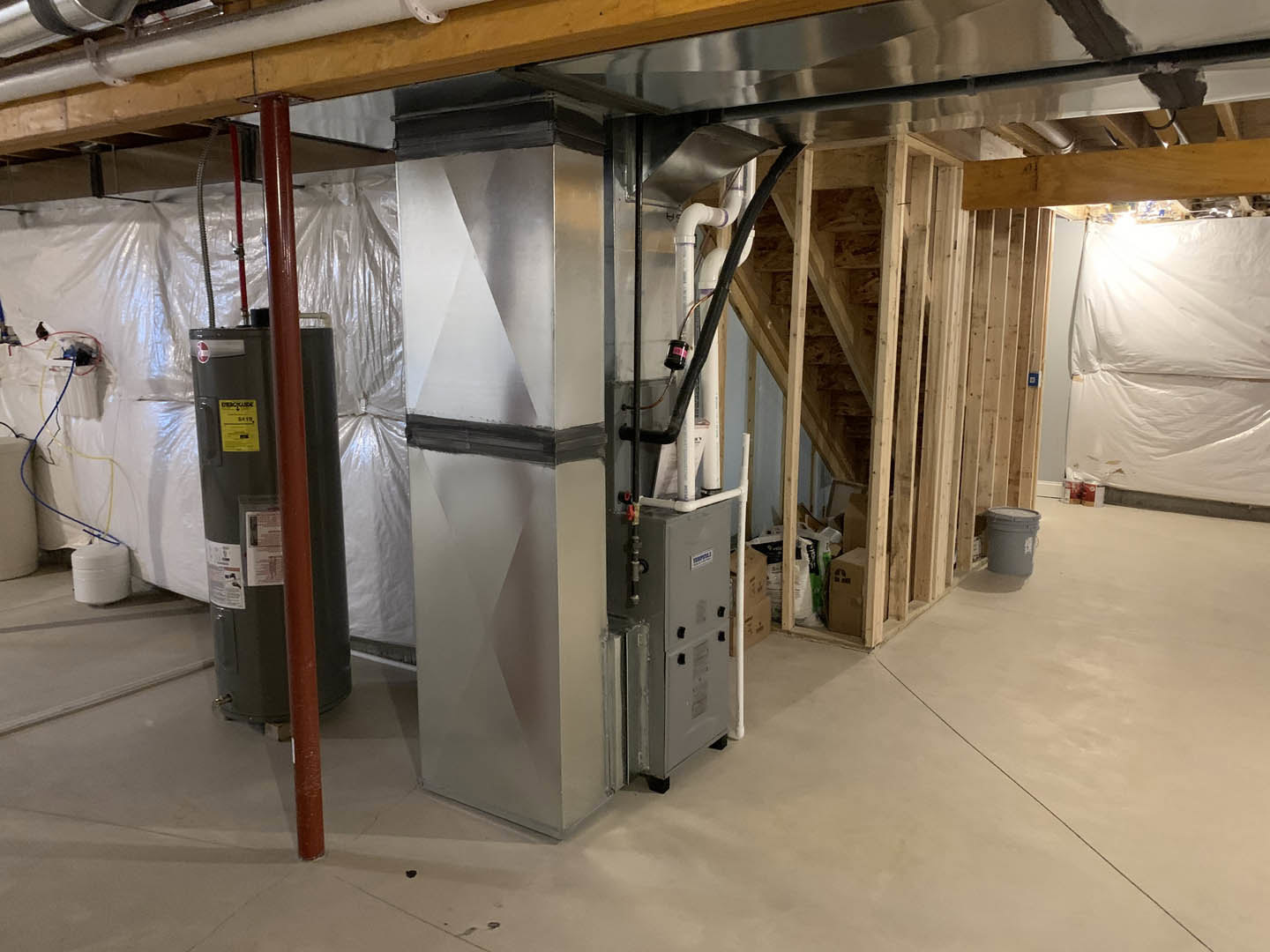 Basement room with exposed steel beam ceiling, grey metal box, black cylinder with white labels, white plastic sheet covering part of wall, white rectangular object secured with