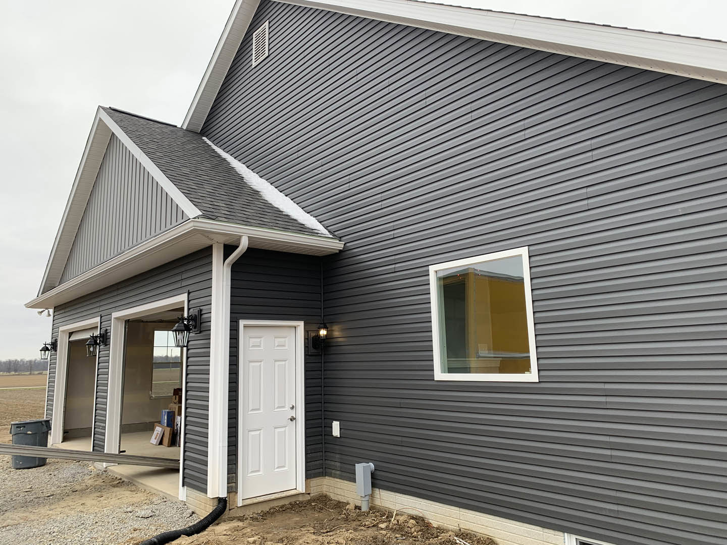 Grey siding house with attached garage, open white entry door with silver handle, white-framed window, black trash can on dirt driveway, cloudy sky above