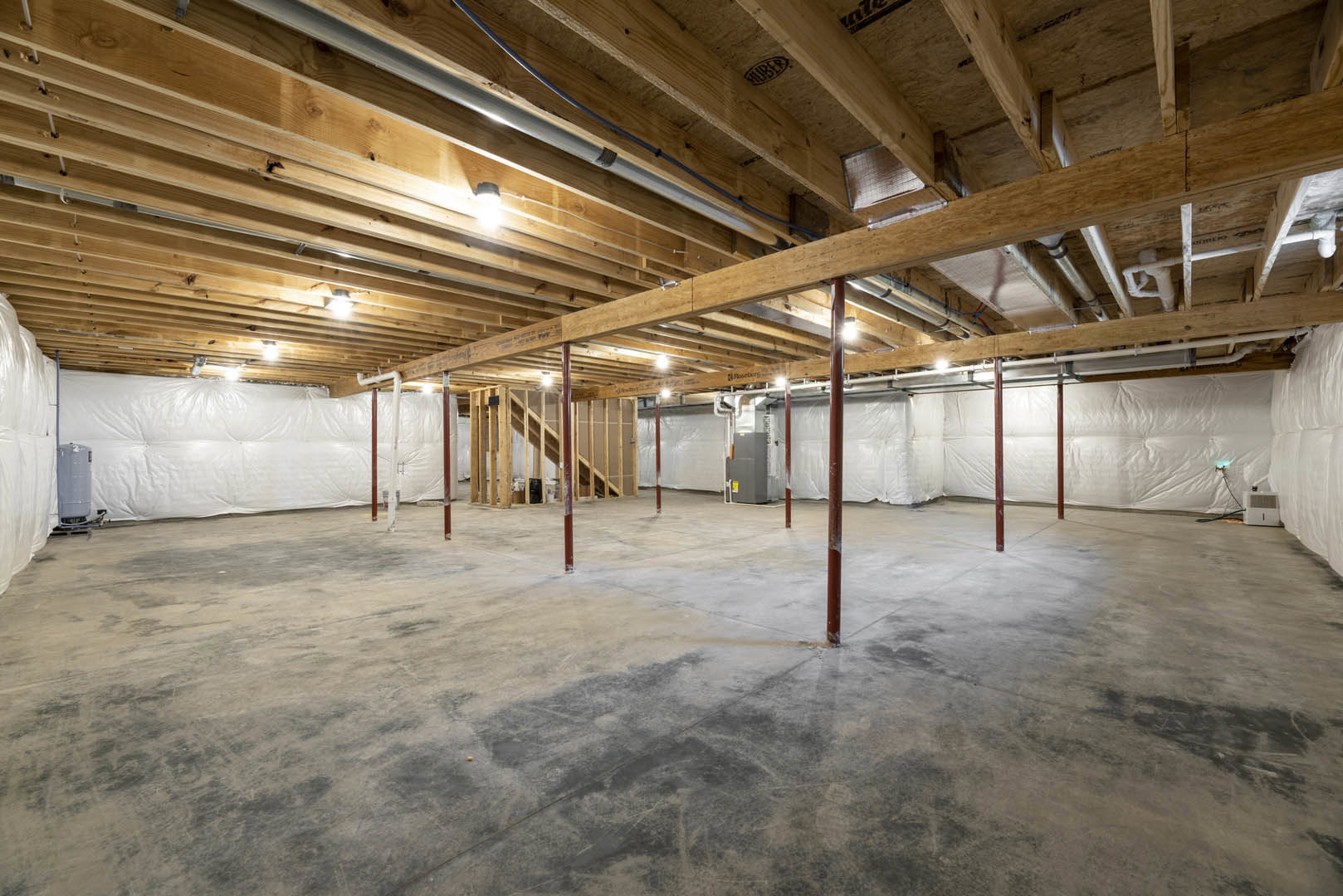 Exposed wooden ceiling beams, white wall with plastic covering, concrete floor with red support poles, unfinished basement interior