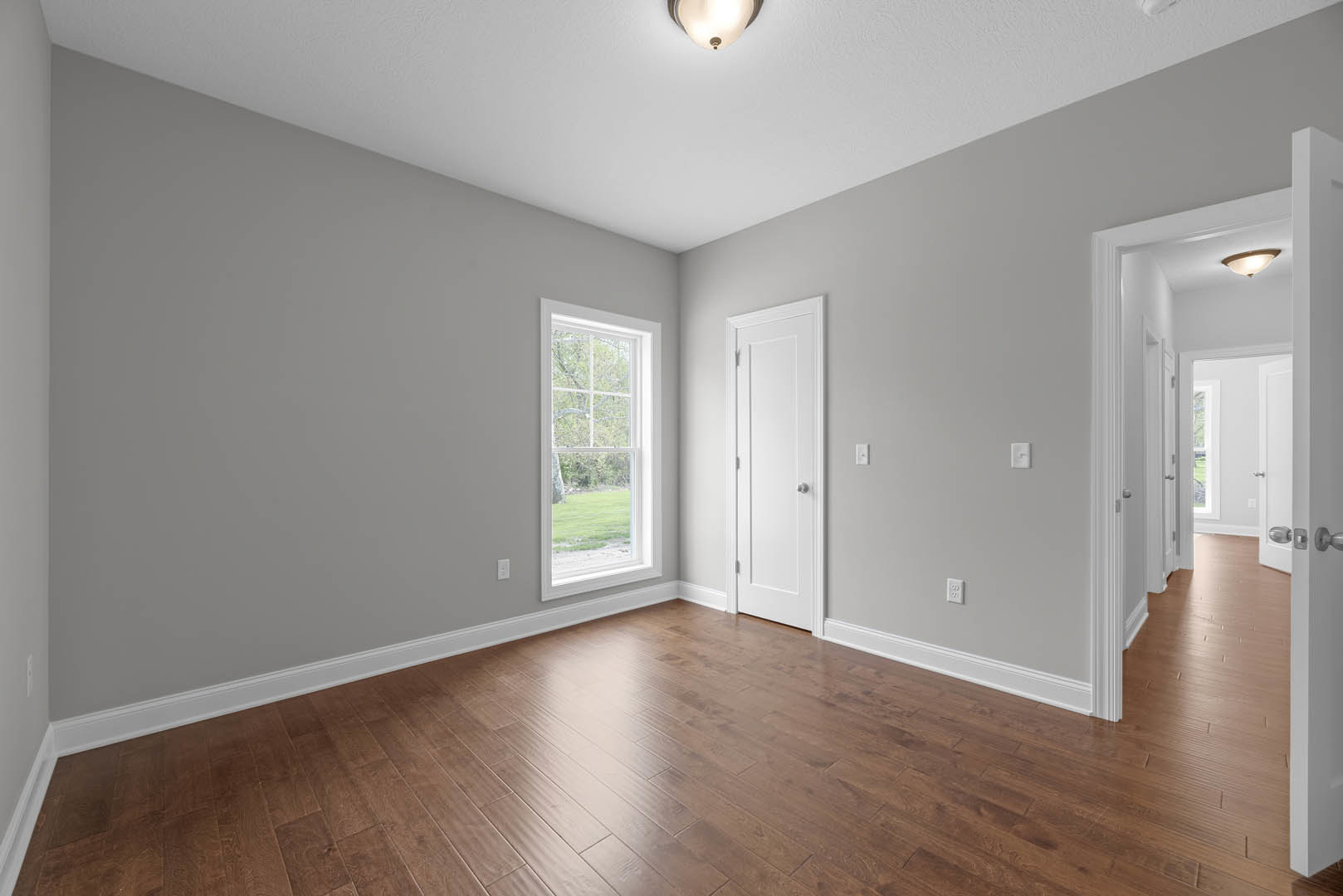 Wood flooring in a room with white plaster walls, a white door with a silver knob, a window overlooking a green lawn, and a ceiling light fixture