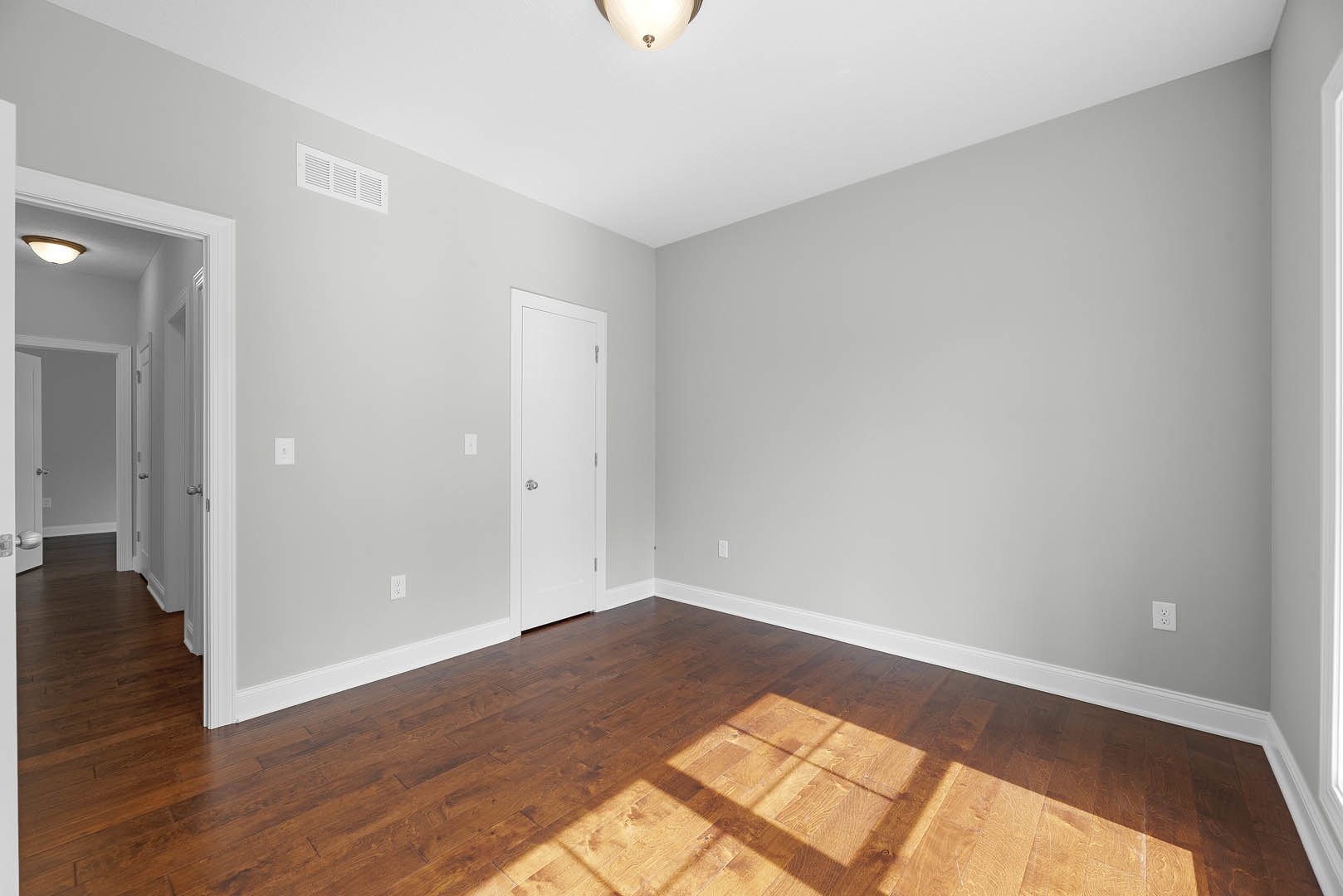 White paneled door with silver handle, wood plank flooring, white walls, ceiling light fixture, wall vent, window with natural light