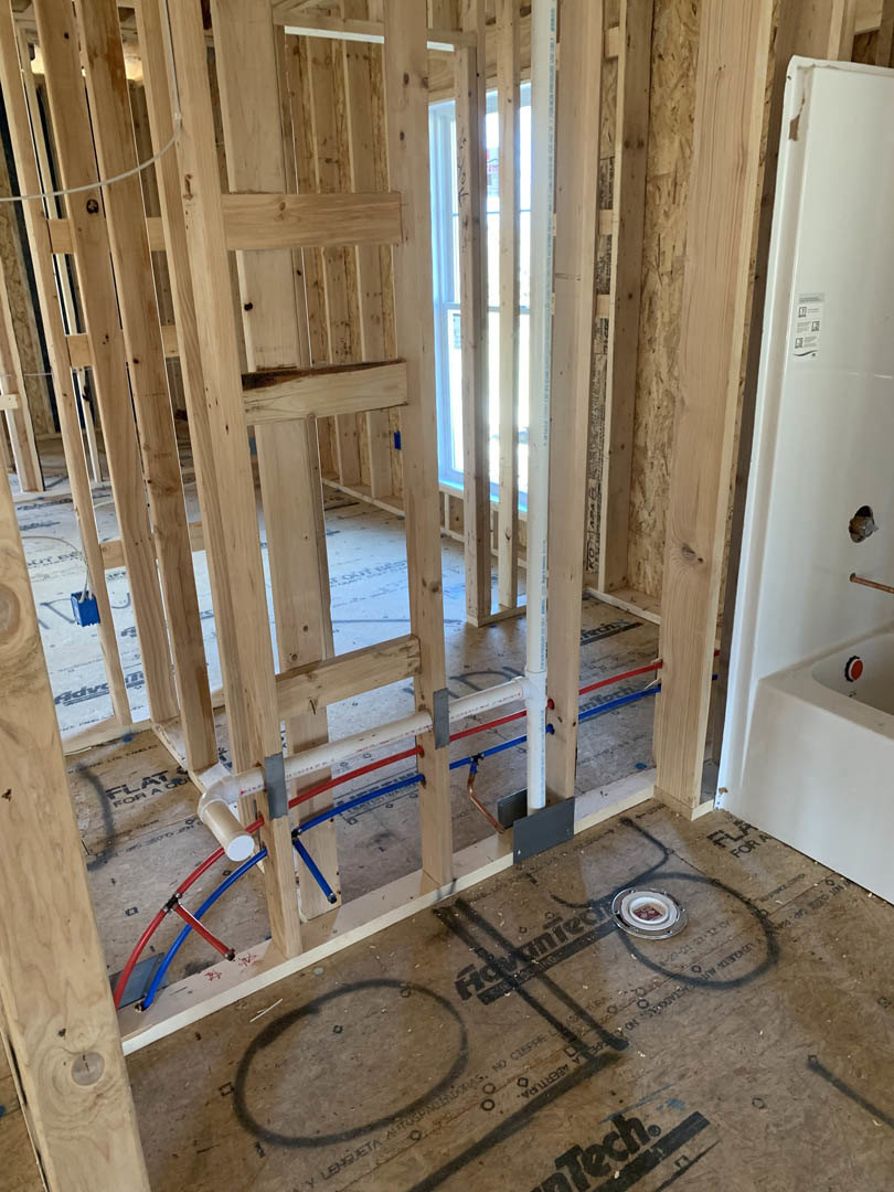 Exposed wood framing and plumbing pipes in unfinished room, open doorway with wall cutout, subfloor with circular hole, construction materials and insulation visible