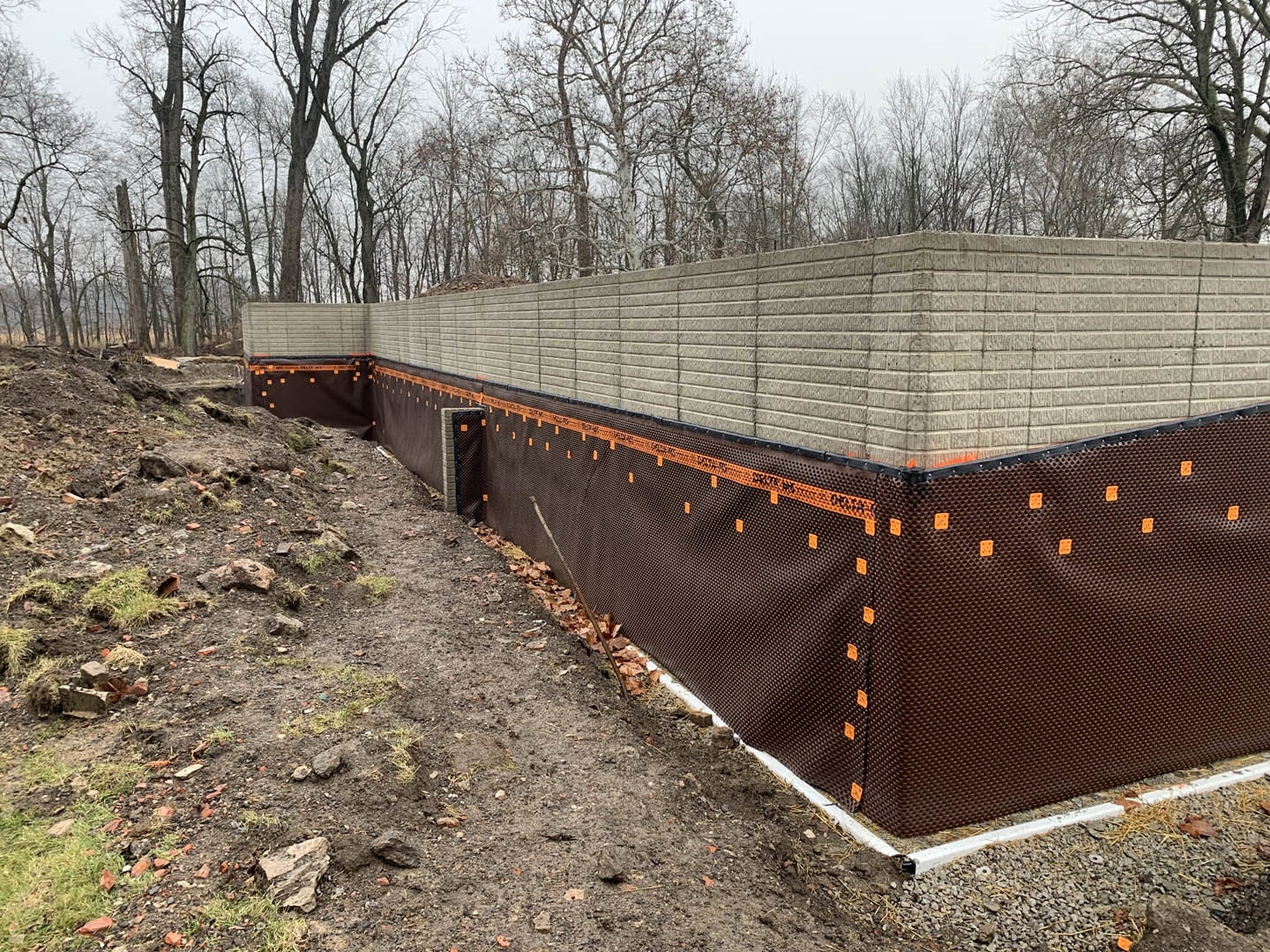 Red brick wall topped with metal fence, orange construction tape and signs attached, trees and sky visible in background.