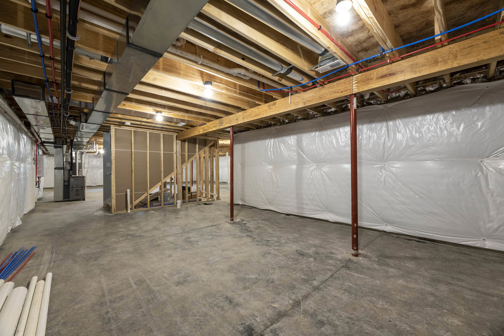 White walls and exposed wooden ceiling beams above a room with unfinished wooden floors partially covered by white plastic sheeting, visible staircase with wood paneling, and white