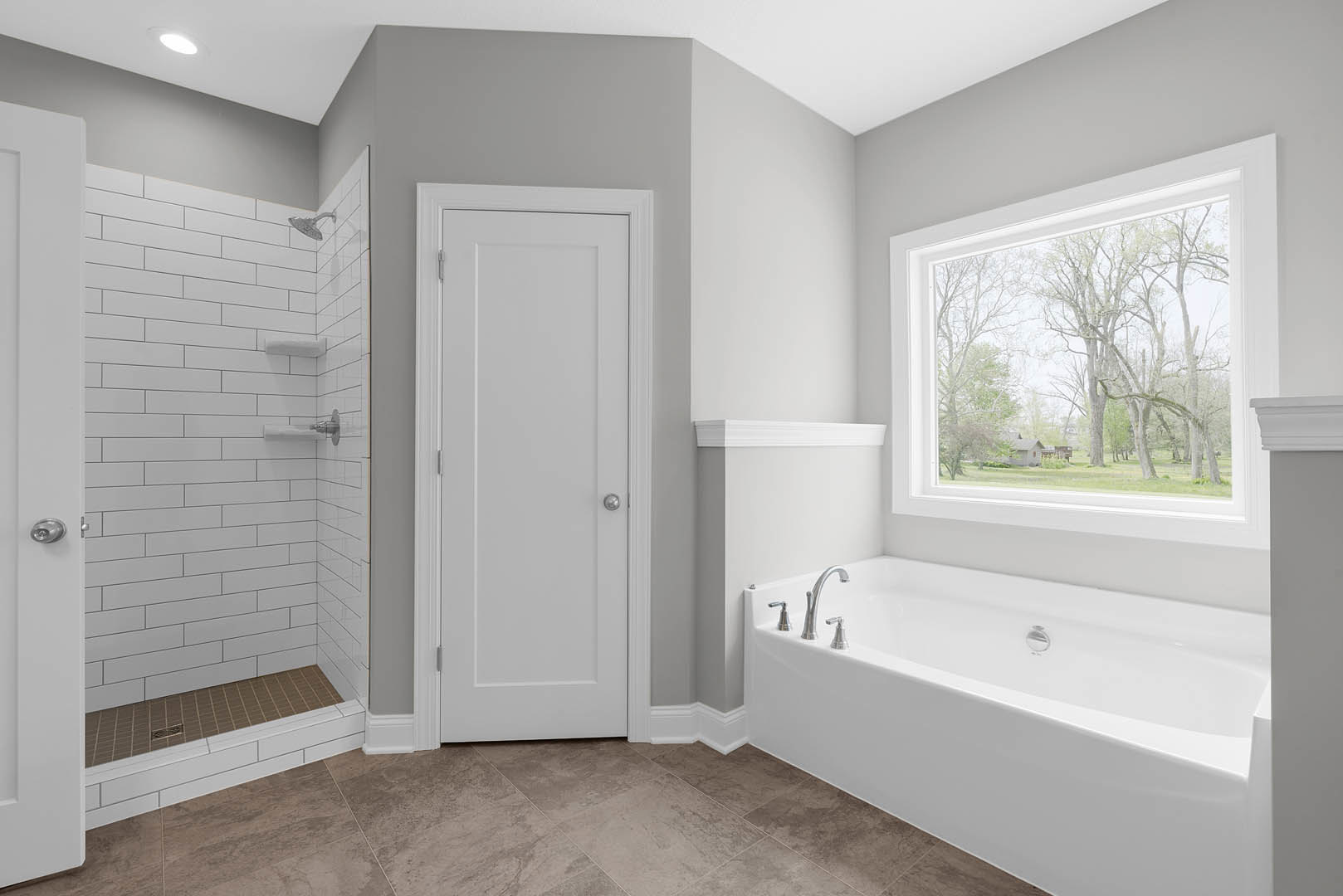Bathroom with white tile shower featuring metal shelf, built-in bathtub with chrome faucet, white door with silver round knob, window overlooking trees, light-colored walls and