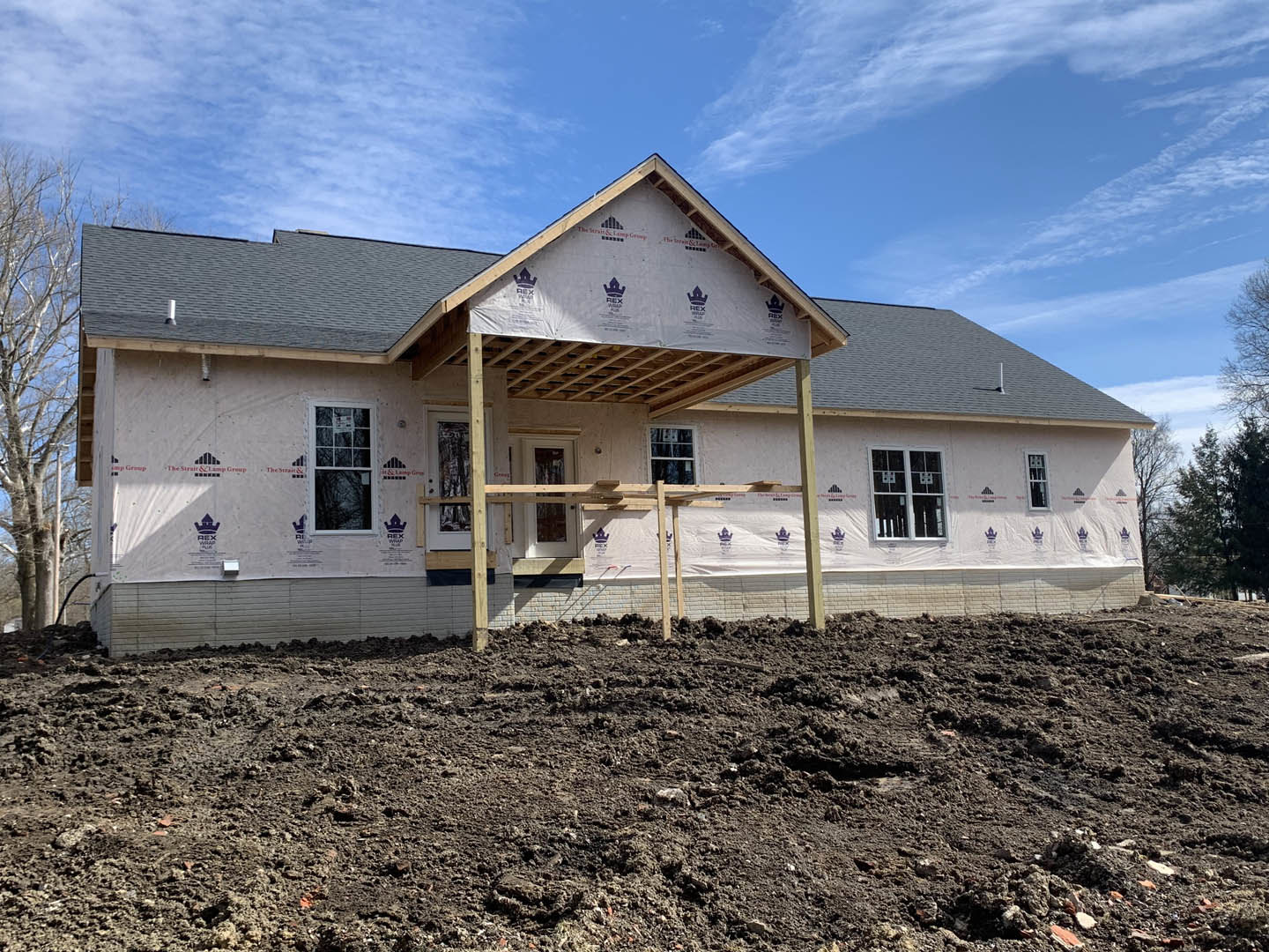 Two-story house under construction with exposed wood framing, white window frames, wooden porch, muddy ground, and temporary fencing in a dirt field.