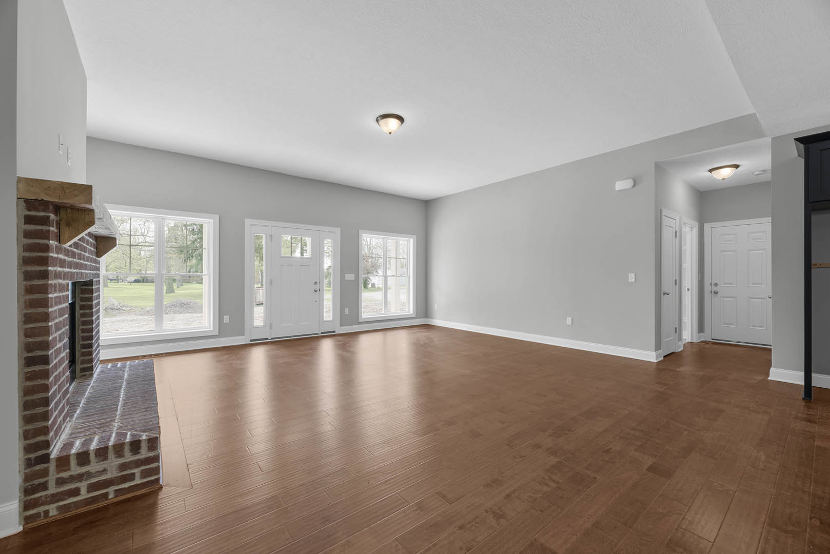 Living room with wood flooring, white fireplace, large window overlooking yard, white door with glass panes, ceiling light fixture, plaster walls.