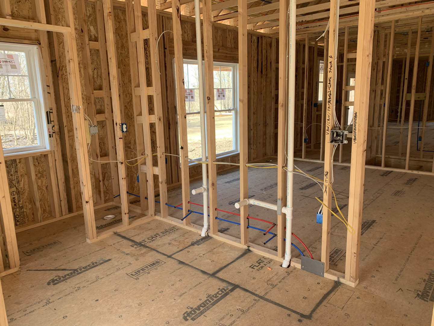 Unfinished room with exposed wooden framing, visible pipes and electrical wiring, window with a permit sign, construction materials scattered on plywood subfloor