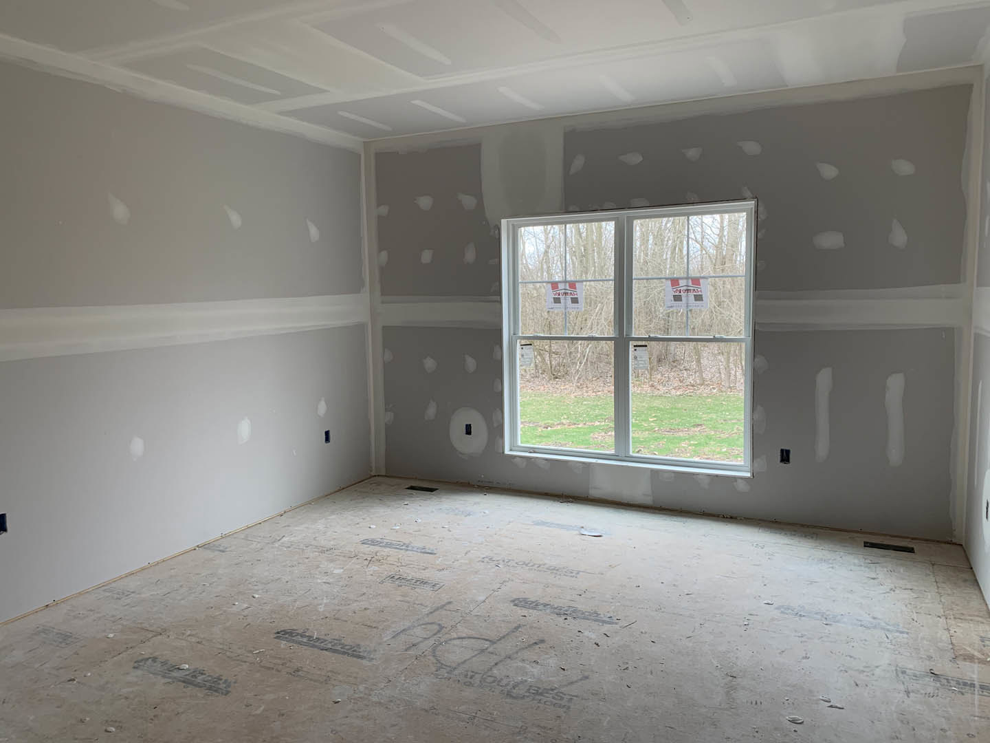 Bright room with white plaster walls, two large windows, light-colored flooring, and red and white signage visible through the window.