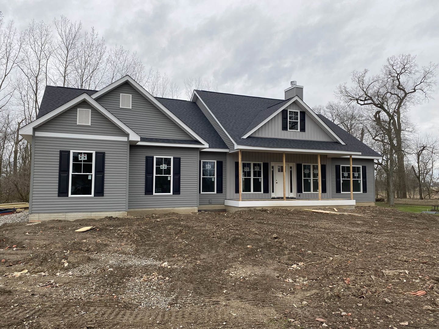 Partially built house with grey roof, white framed windows, white wall vent, basketball hoop, exposed dirt patch, surrounding trees, and cloudy sky.