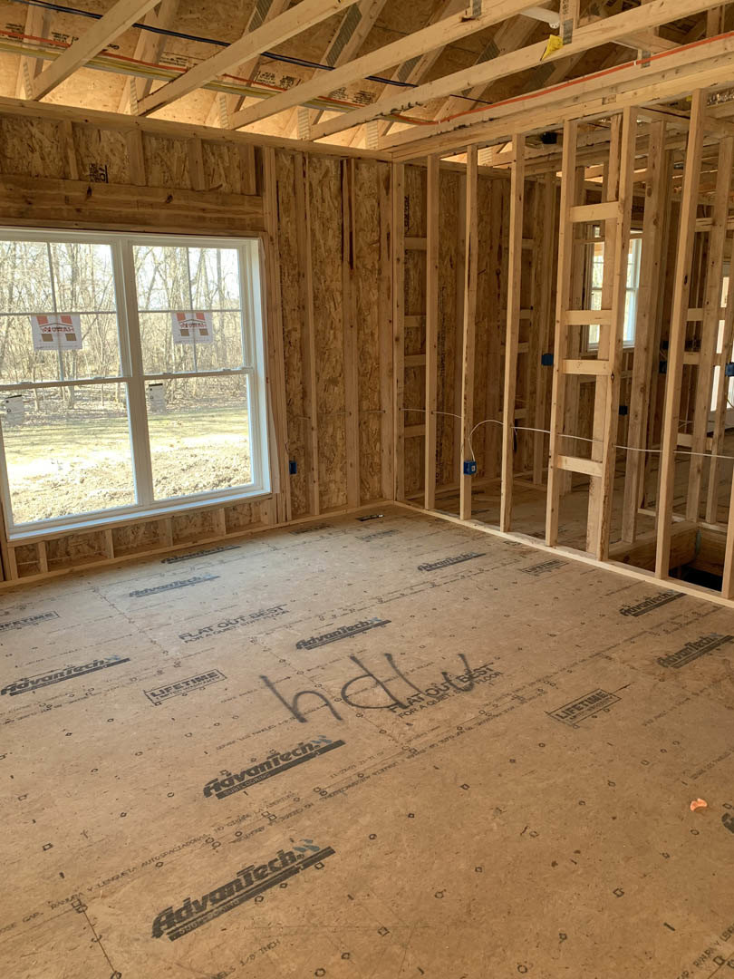 Room under construction with exposed wood framing, multiple windows, unfinished floor, and insulation visible between beams; red signage and logo stickers on window glass.