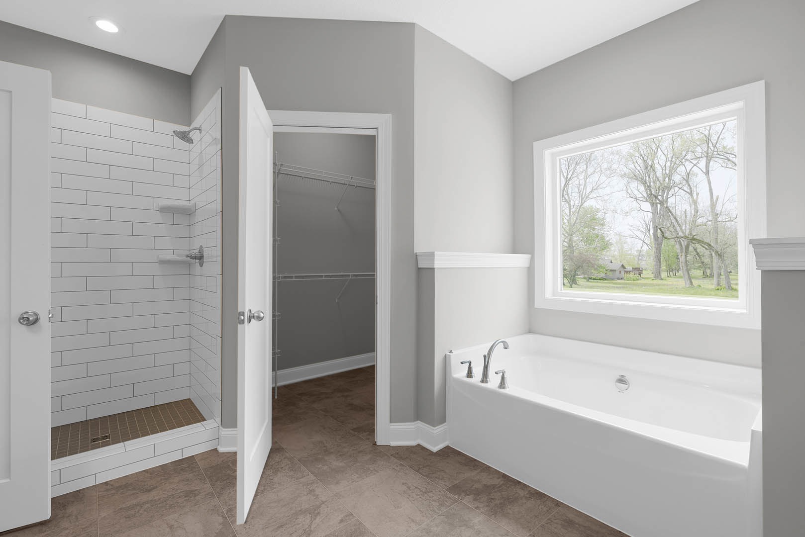 White tile bathroom featuring a freestanding bathtub with silver faucets, glass-enclosed shower with metal shelf, large window overlooking trees, and visible door handle and