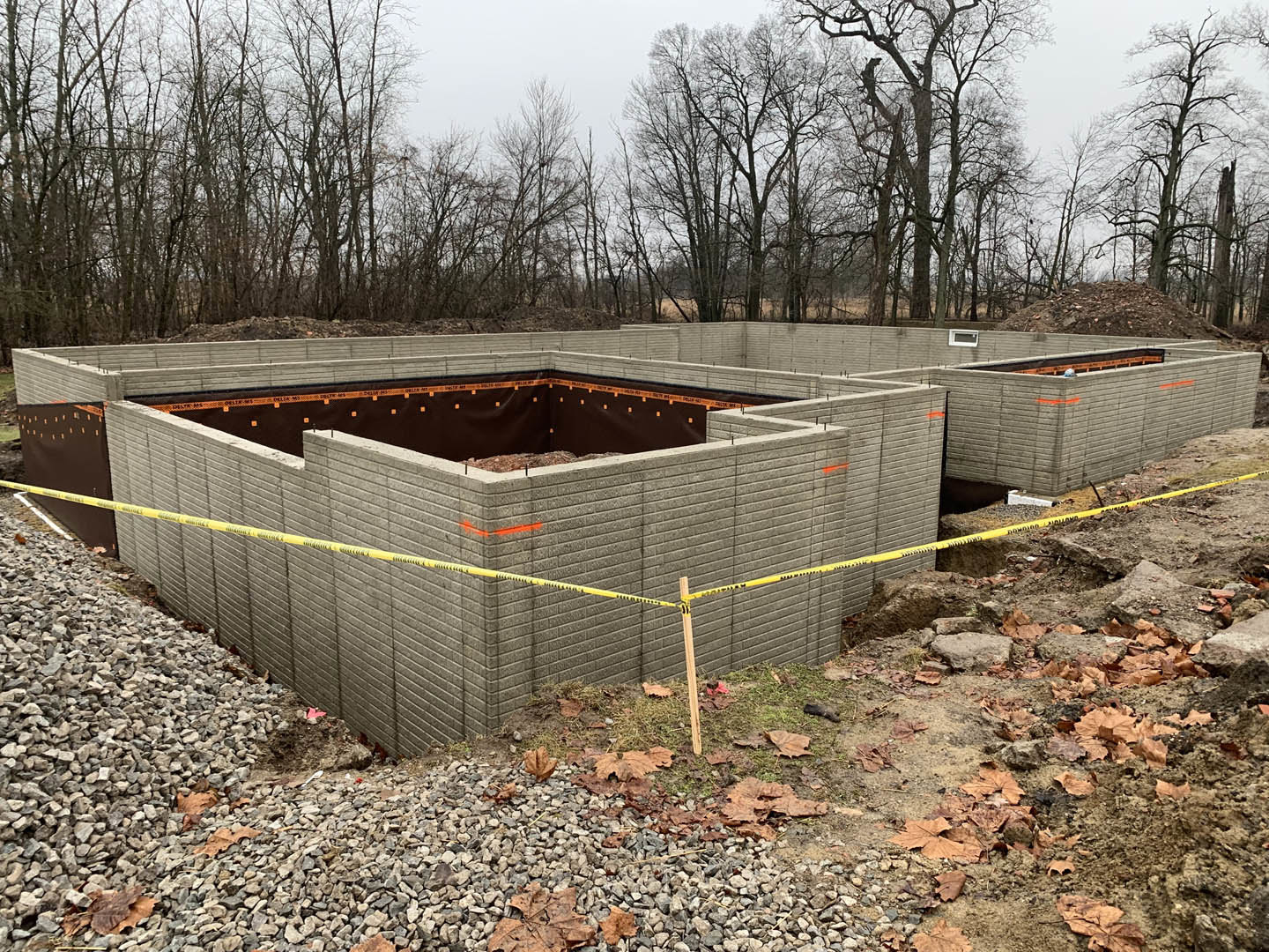 Partially built home with exposed concrete foundation, yellow caution tape, bare tree, and soil surrounded by temporary fencing