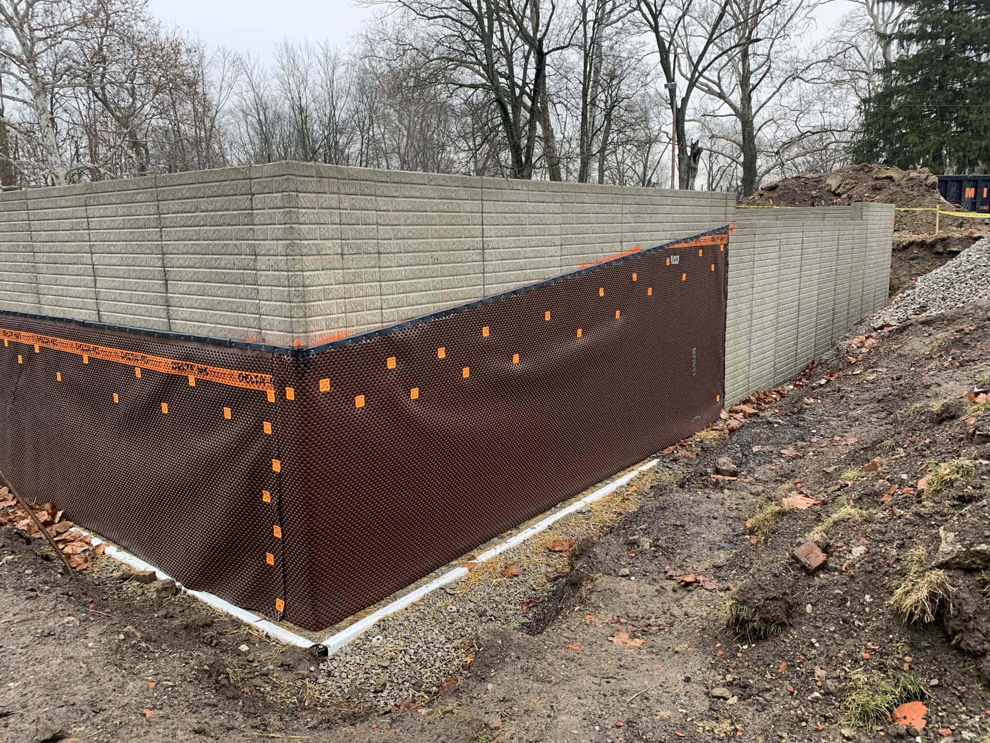 Red brick wall bordered by a metal fence, brown tarp draped over part of the wall, black container with orange lettering nearby, snow-covered trees in background, concrete block