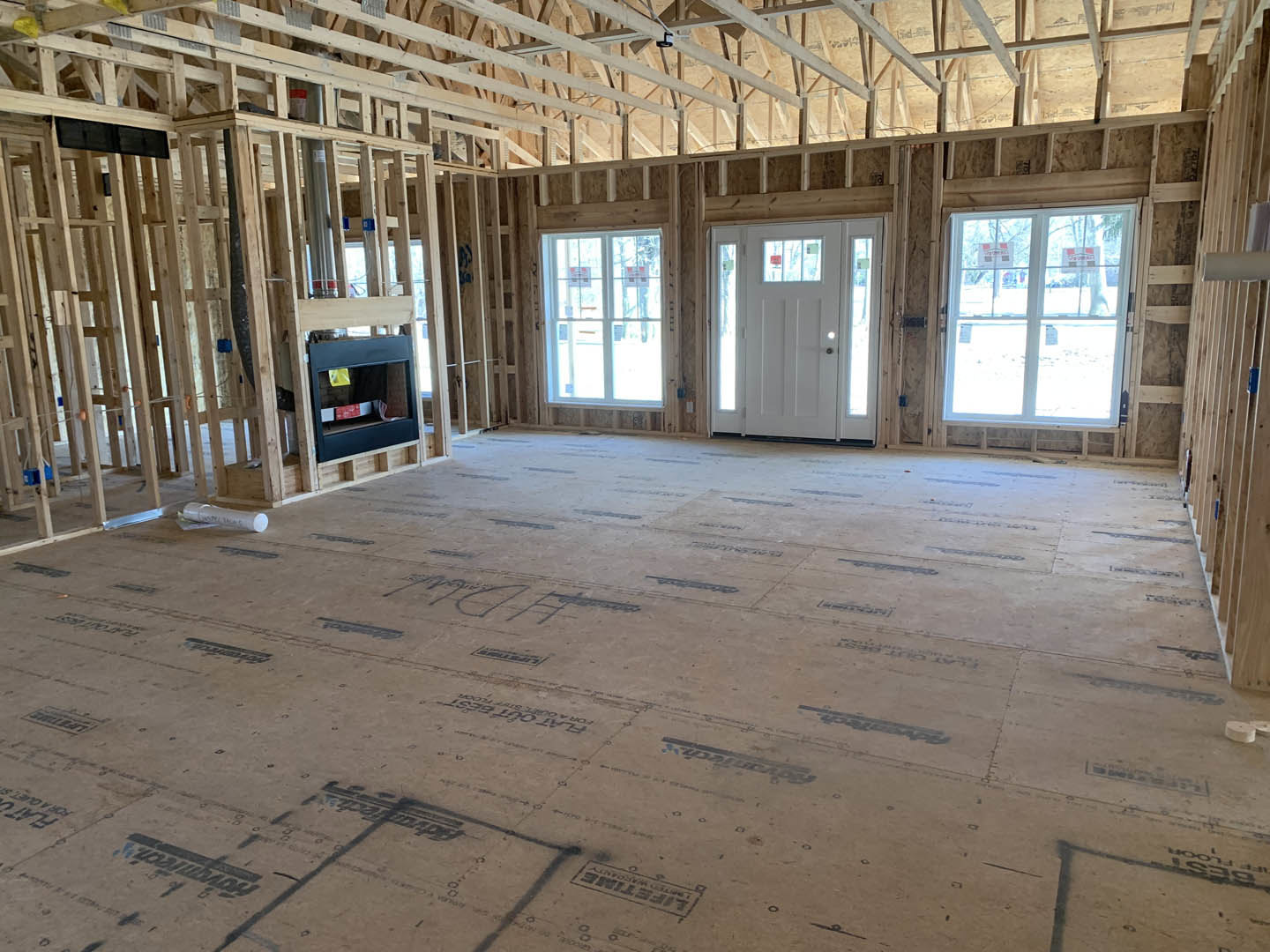 Living room with exposed wood ceiling beams, stone fireplace, white glass-paneled door, window displaying signs, and unfinished floor marked with black writing