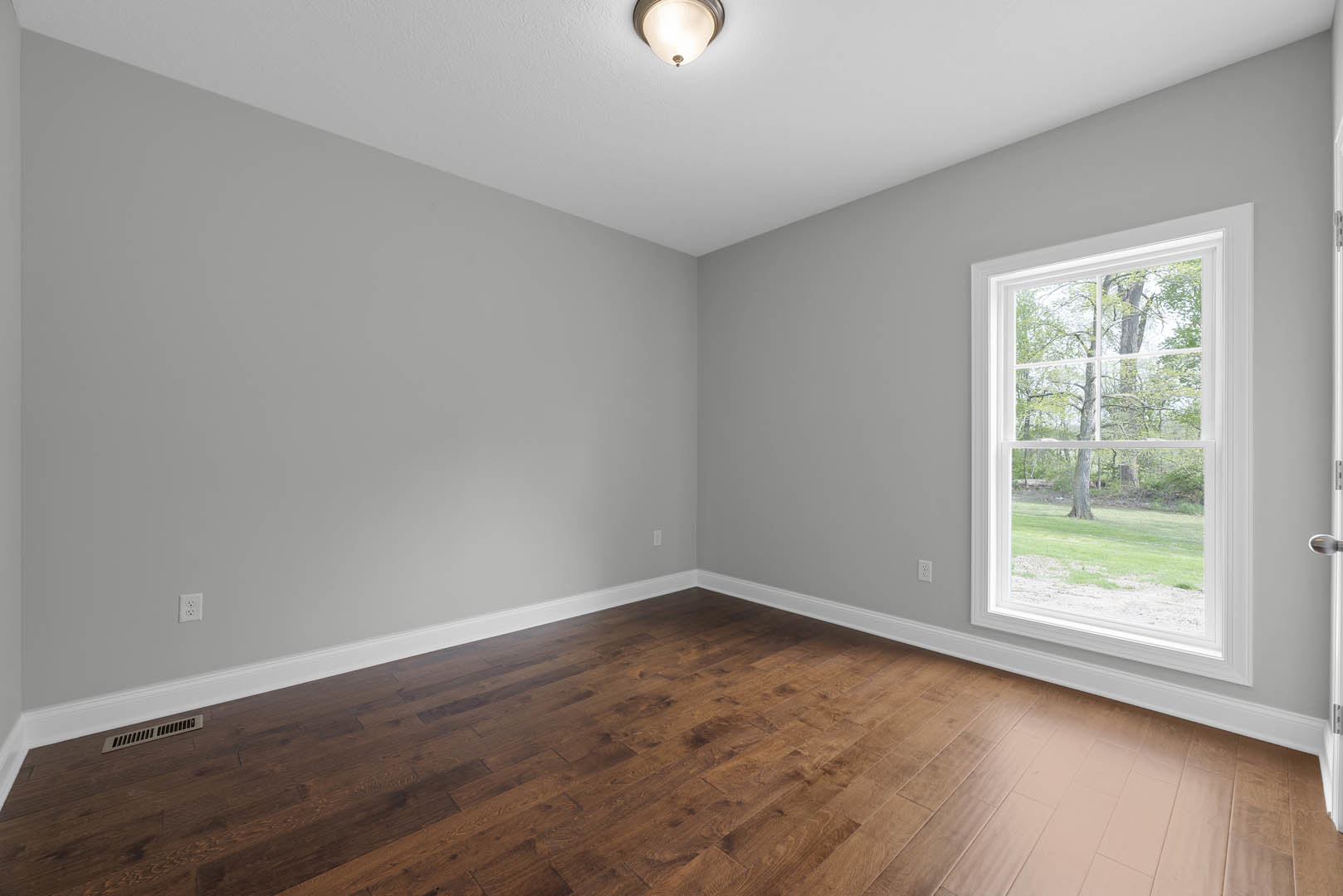 Hardwood floor with white baseboards, large window overlooking trees, round ceiling light fixture, wall vent, smooth plaster walls