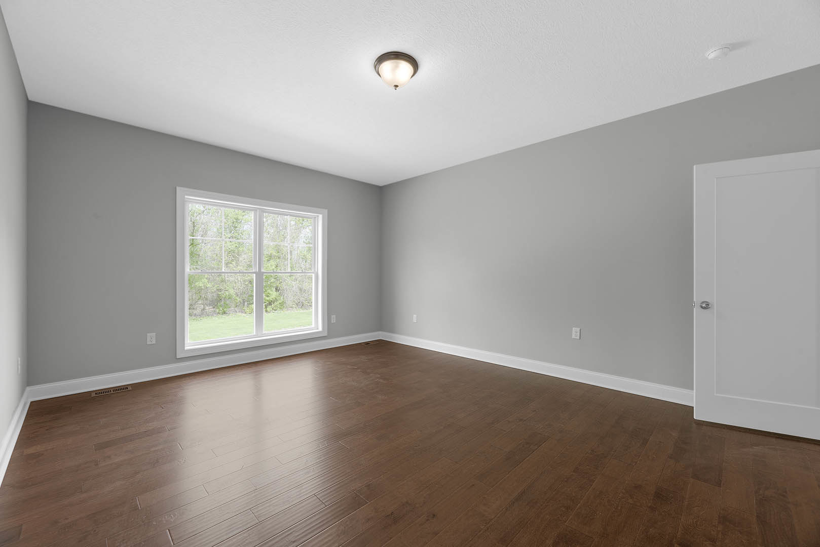 Sunlit room featuring wide-plank hardwood flooring, white walls, large window overlooking leafy trees, recessed ceiling light, and a white paneled door