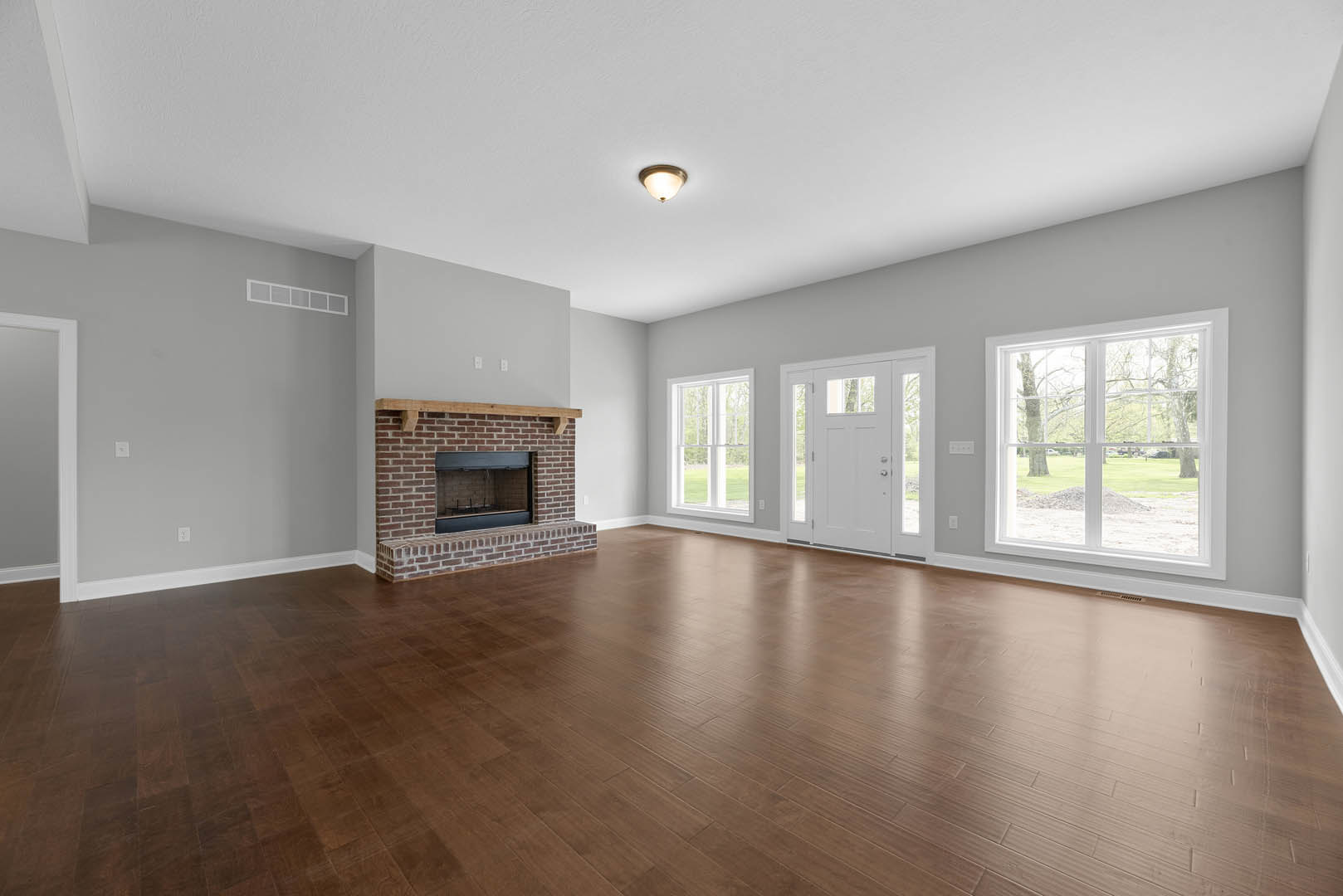 Living room with hardwood floors, brick fireplace topped by a wood mantel, large window overlooking green lawn, and ceiling light fixture.