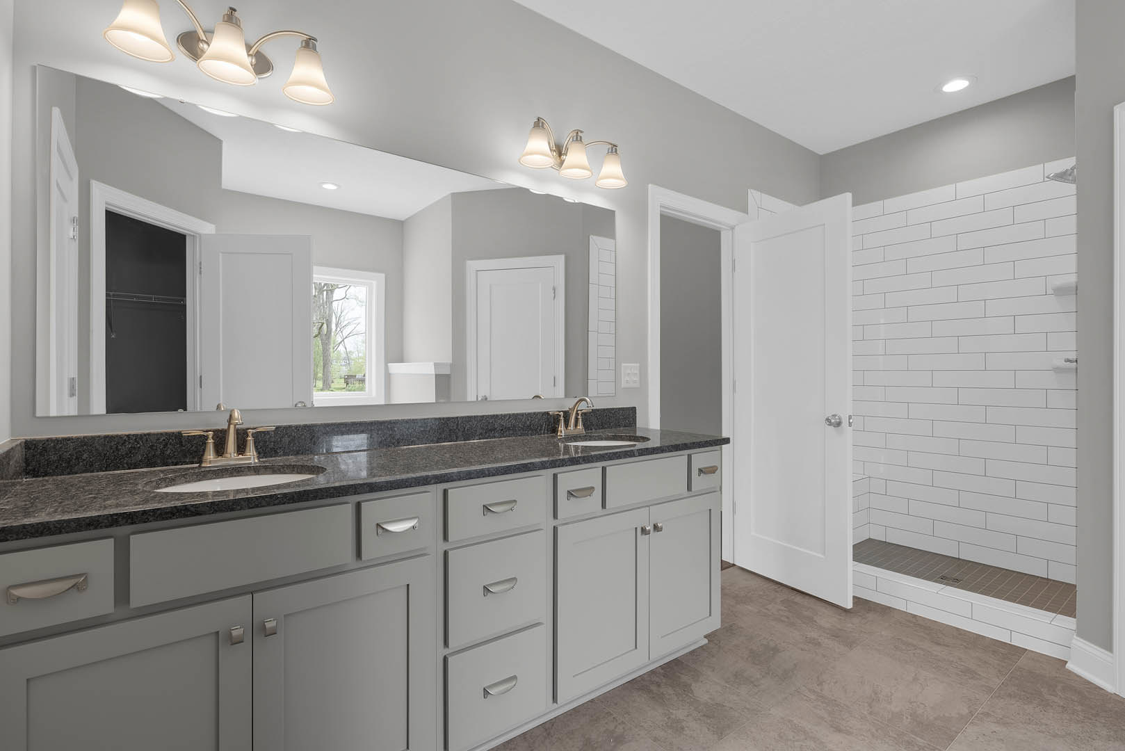 Bathroom featuring a double sink vanity with black countertop, white cabinetry, glass-enclosed shower, three-light fixture above mirror, window with tree view, and tiled walls.