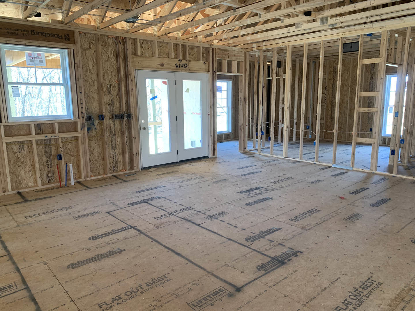 Open room with exposed wood framing, double glass-panel doors, window displaying a sign, unfinished floor marked with construction drawings, and visible ceiling beams