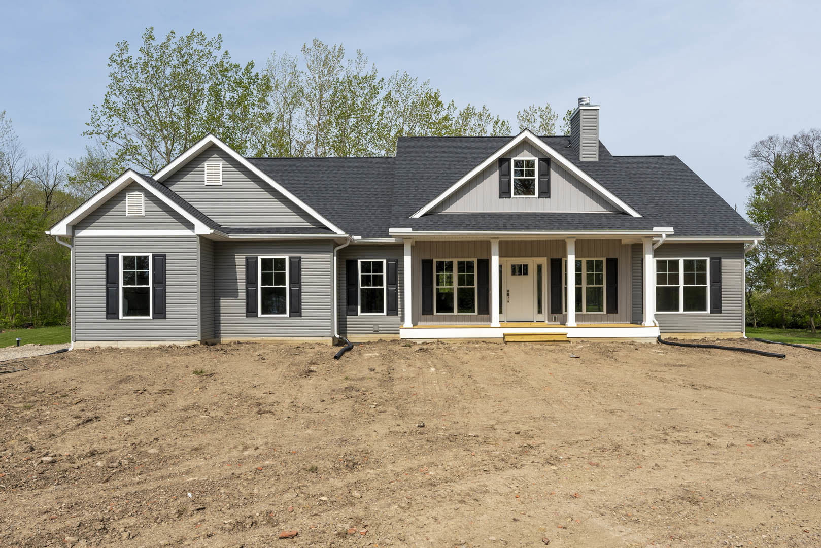 Two-story house with white siding, black shuttered windows, glass-paneled front door, covered porch, and dirt yard with exposed pipe.