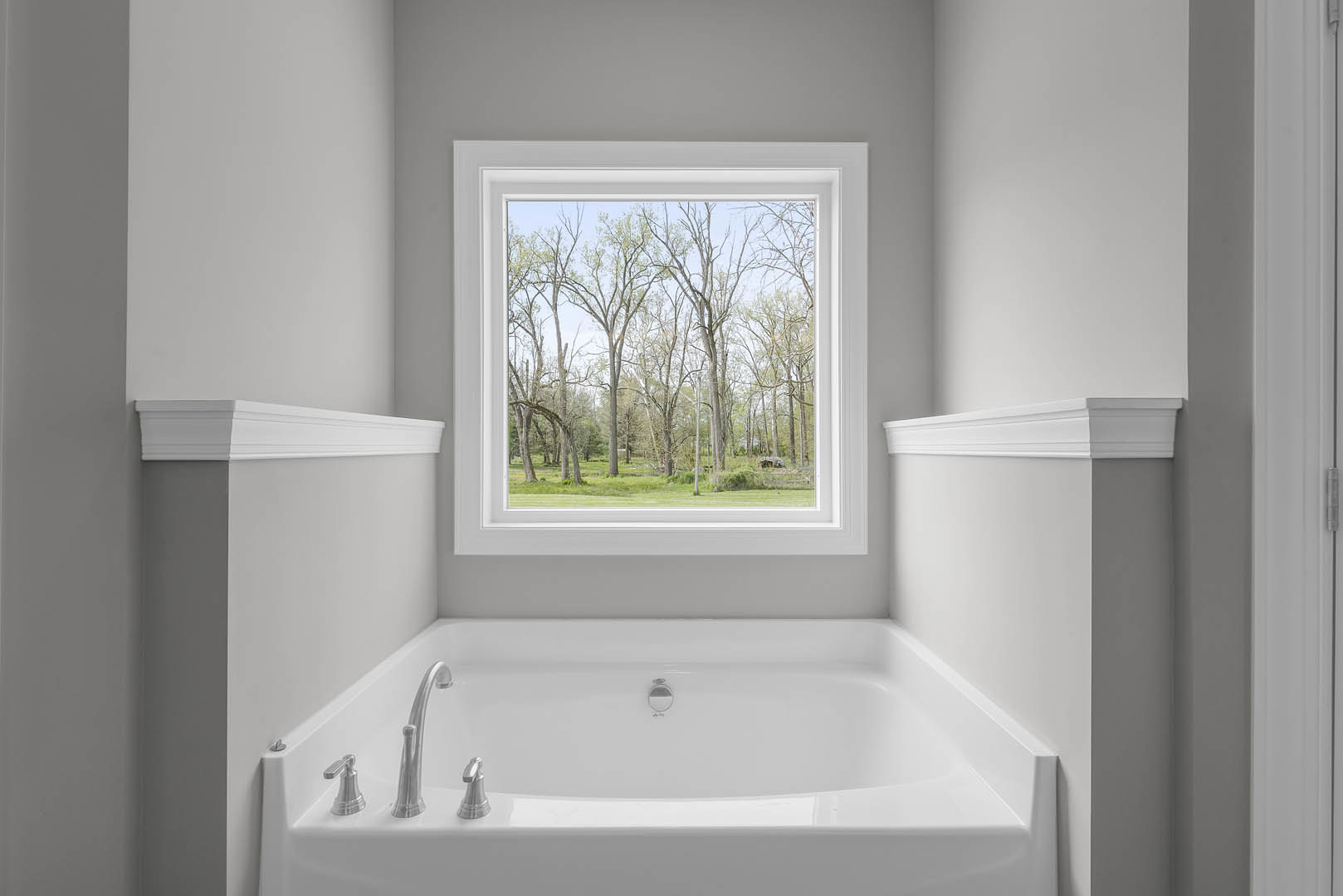 Freestanding white bathtub beneath large window with leafy trees visible outside, surrounded by white walls and chrome fixtures