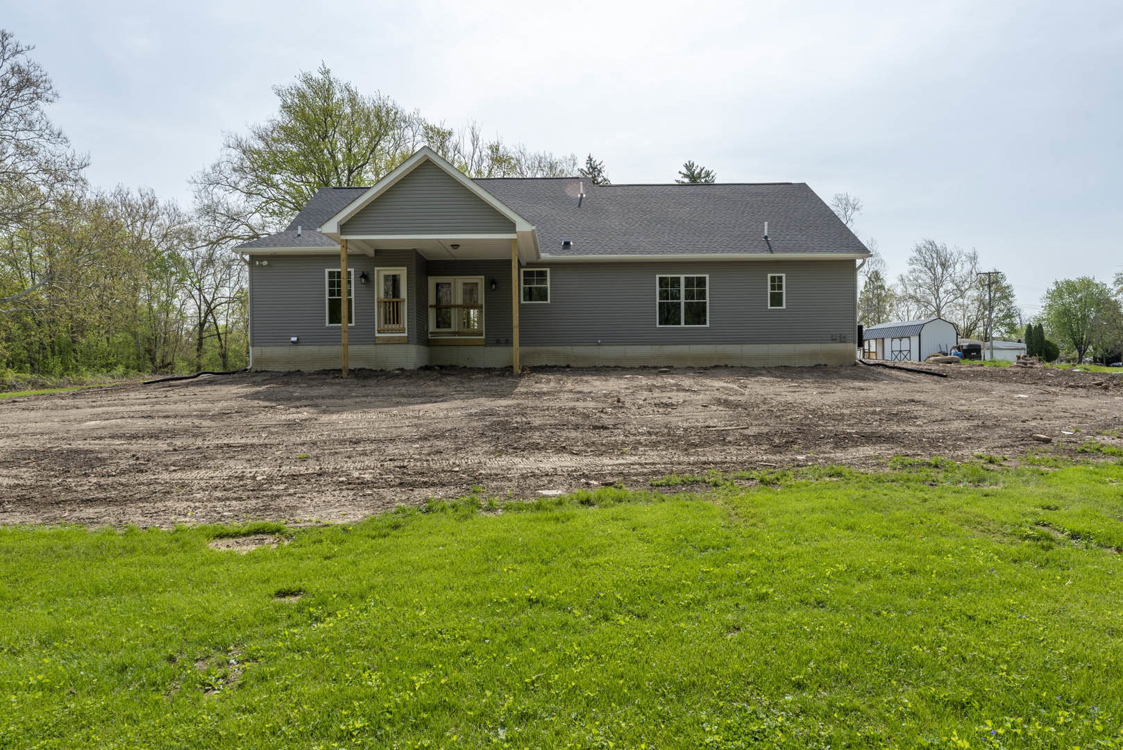 Modern farmhouse with white-framed windows, dirt yard in front, small tree near the entry, grassy field beside a dirt road, clear sky overhead